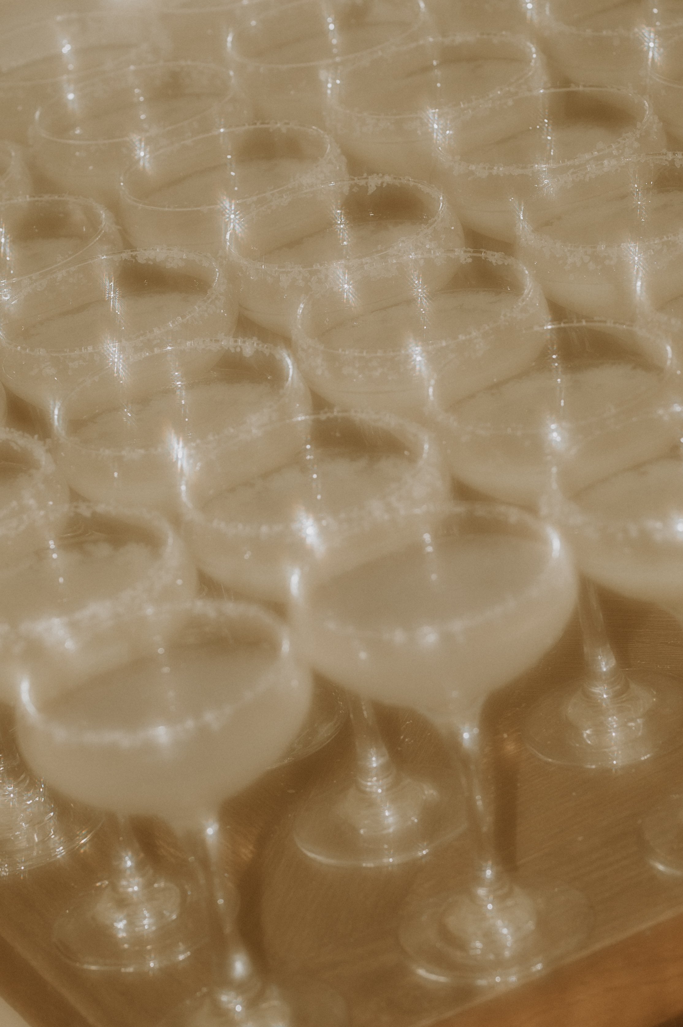 Rows of cocktail glasses with salted rims, filled with a light-colored liquid, likely margaritas, on a wooden surface.