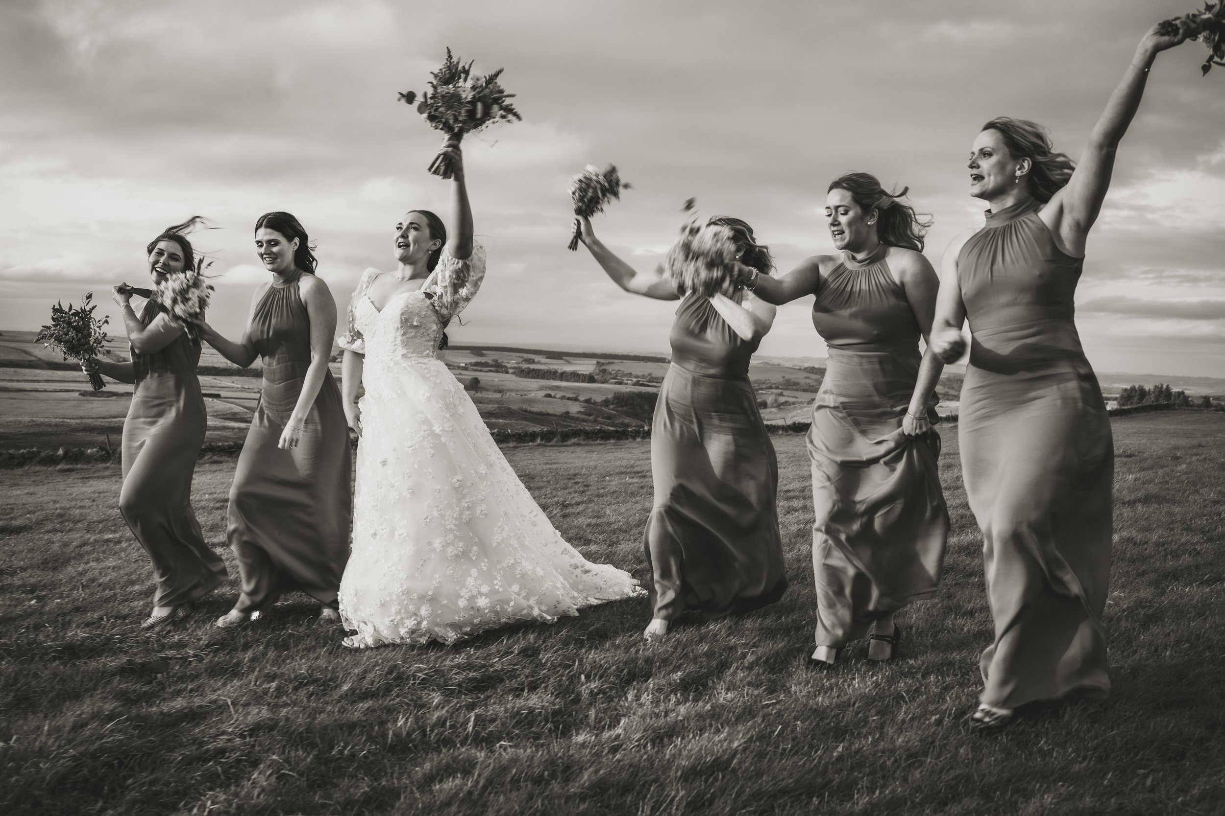 Bride and bridesmaids celebrating outdoors, holding bouquets, with a scenic landscape in the background.