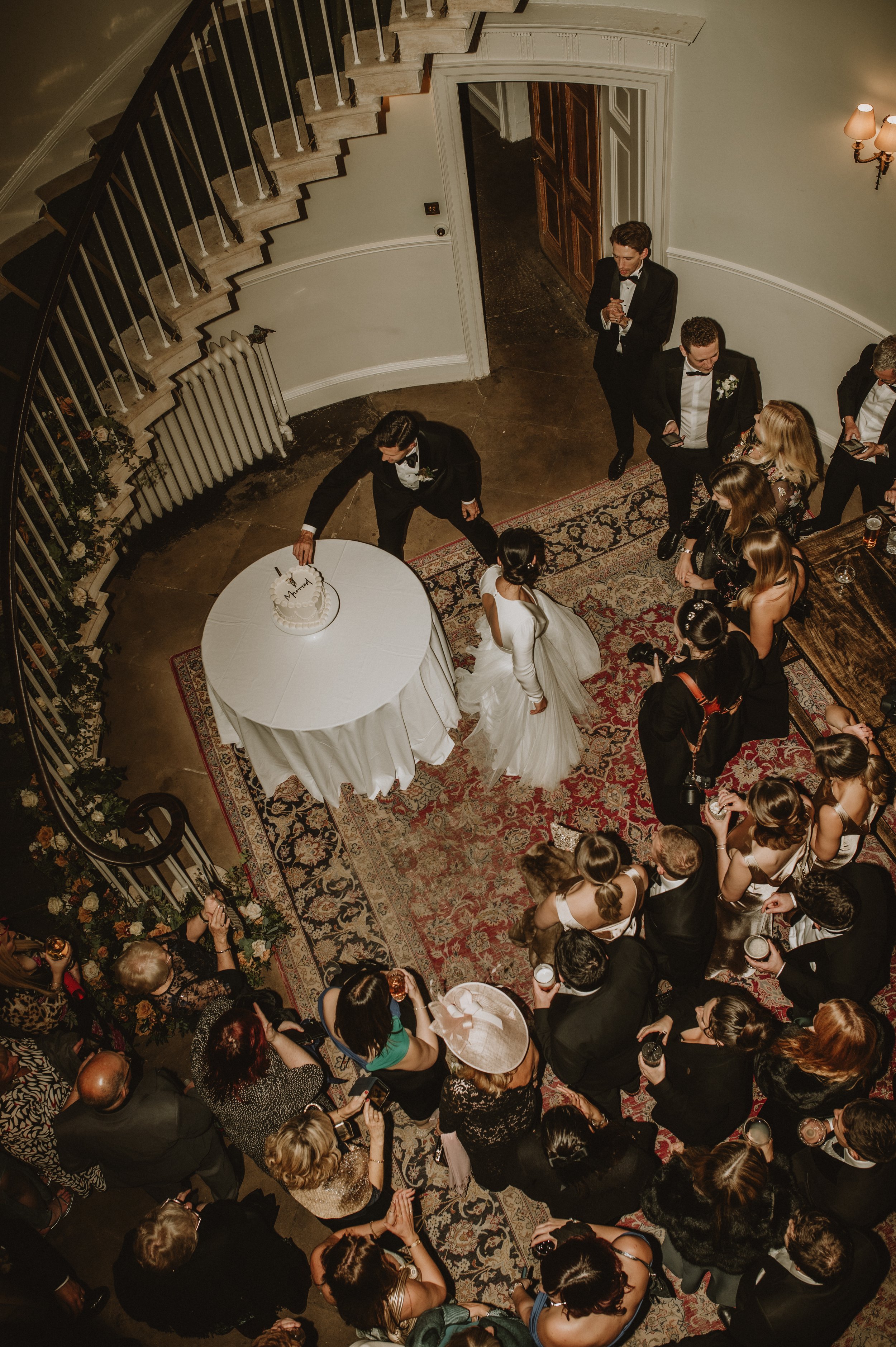 Overhead view of a wedding scene with a couple in formal attire near a cake on a table, surrounded by guests on a patterned rug under a spiral staircase.