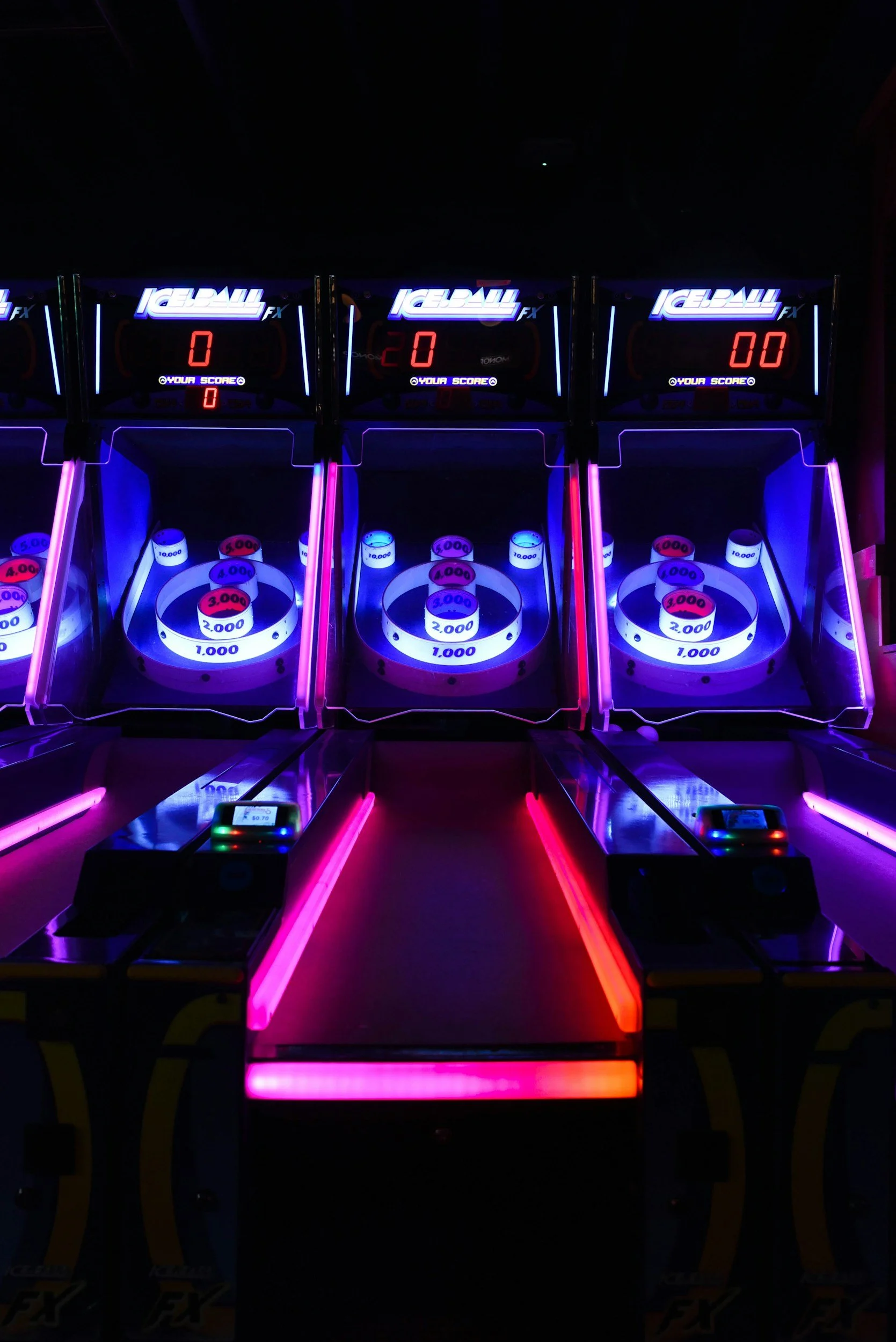 Dark room with three neon-lit pinball machines labeled 'Ice Ball FX' and digital scores displayed but all showing zero. The machines have glowing pins and targets, illuminated in purple, blue, and red neon lights.