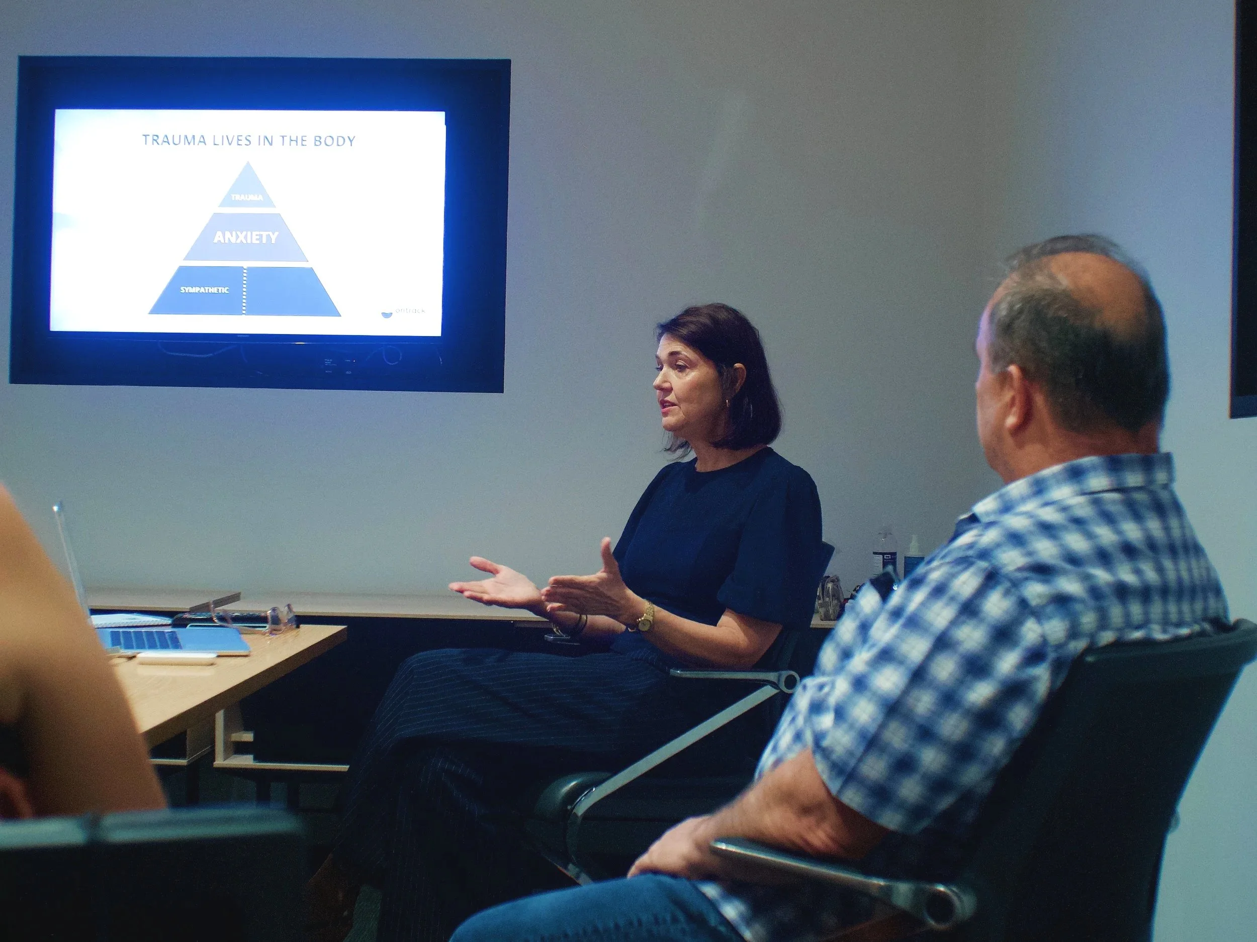 A woman is giving a presentation in a meeting room. A slide titled "Trauma Lives in the Body" with a triangle diagram showing "Sympathetic," "Anxiety," and "Trauma" is displayed on a TV screen behind her. Two men are seated and listening, one in the foreground wearing a checkered shirt.