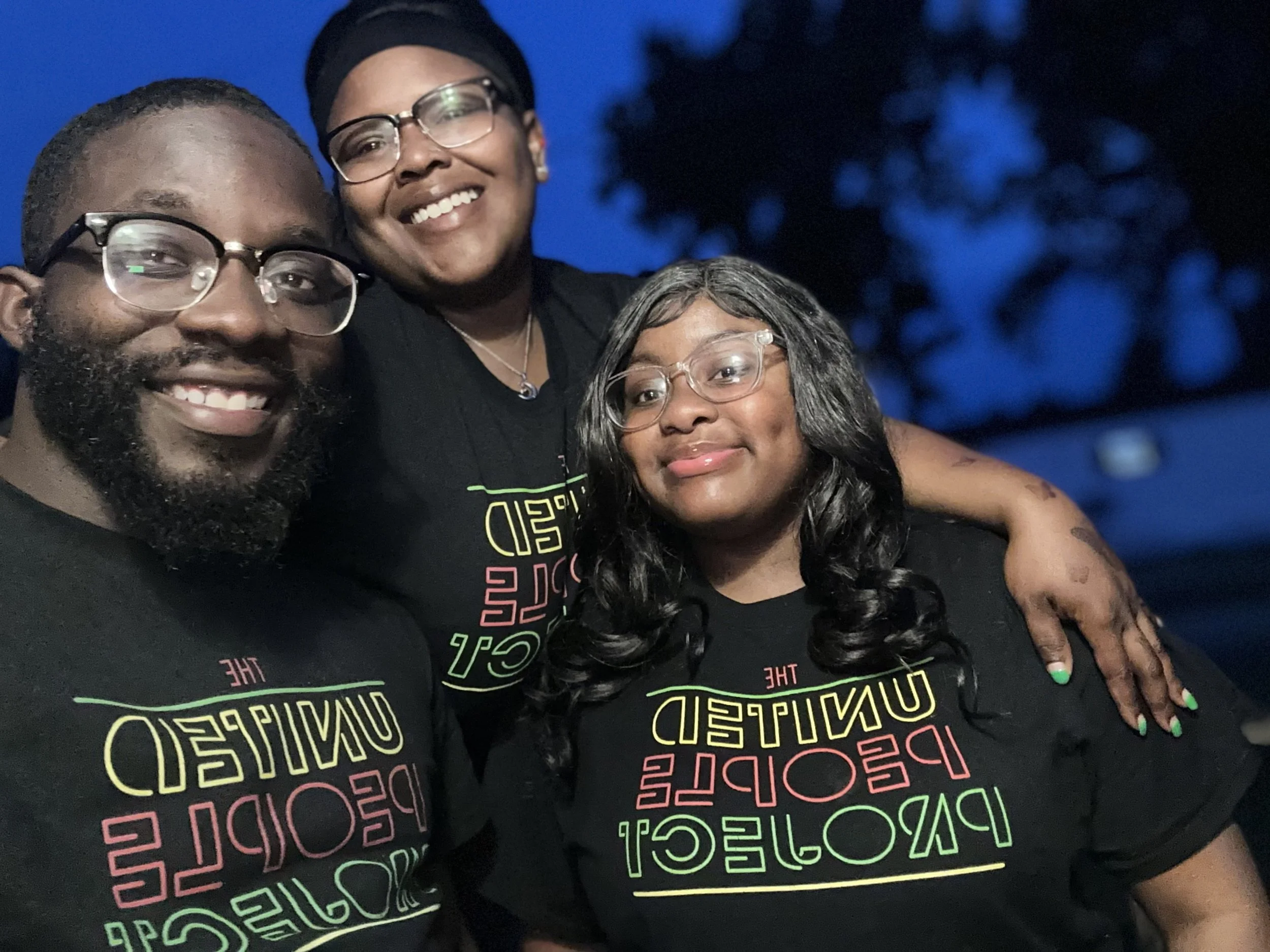 Three people smiling for a photo, wearing matching black T-shirts with colorful text that says 'The United Pedal Project,' standing in front of a dark blue evening sky.