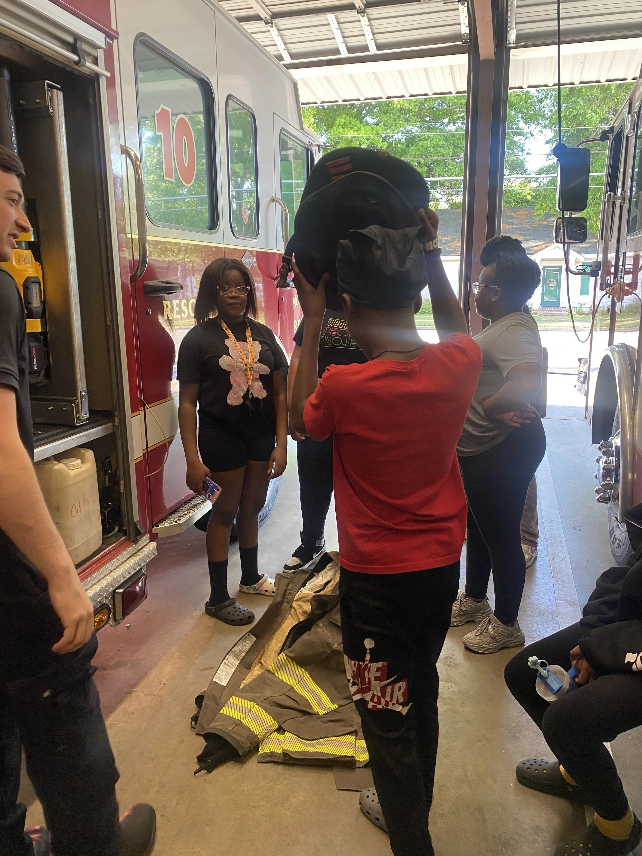 Group of children and adults at a fire station, with a fire truck in the background, participating in a fire safety demonstration.