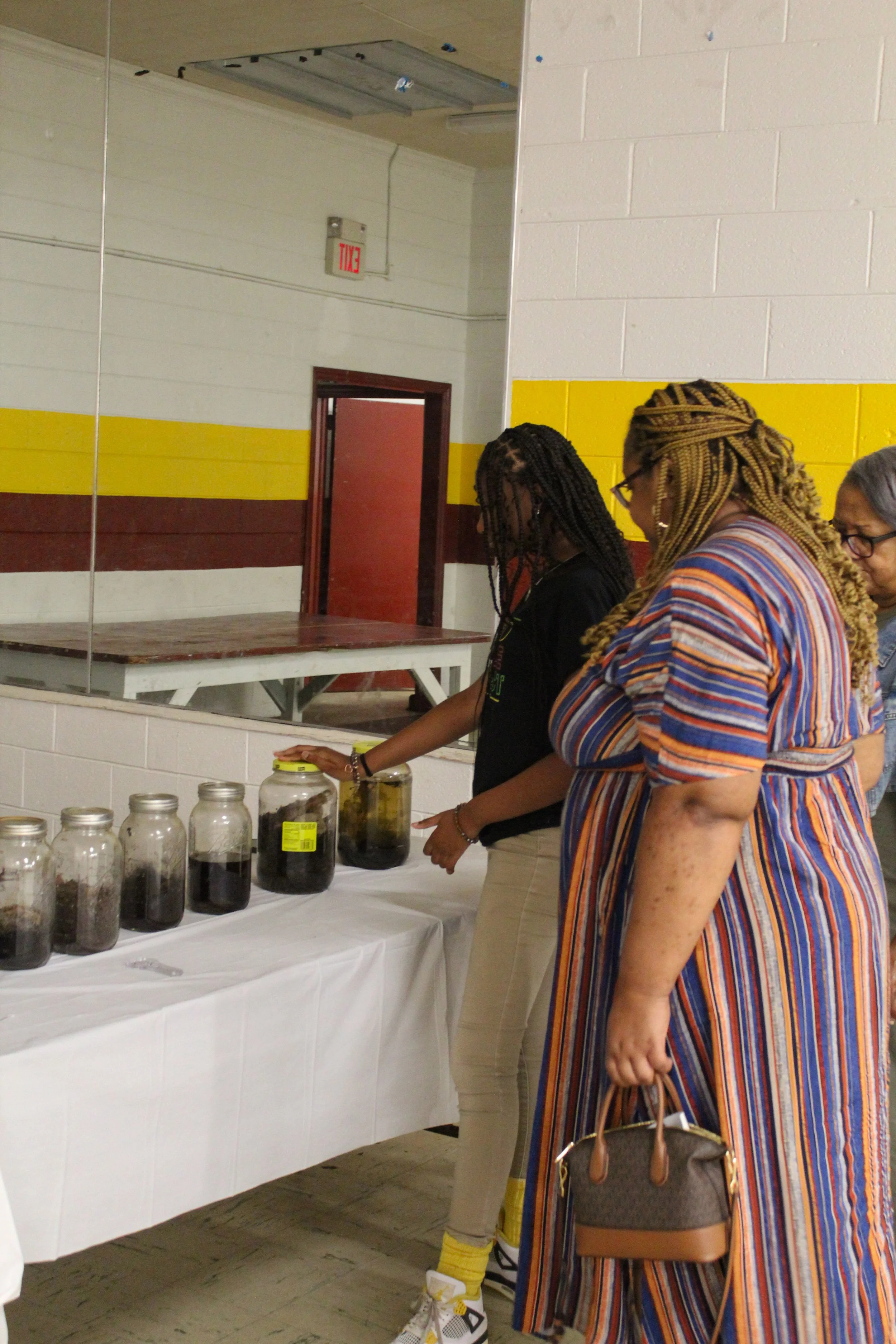 Three women looking at jars with dark contents on a table.