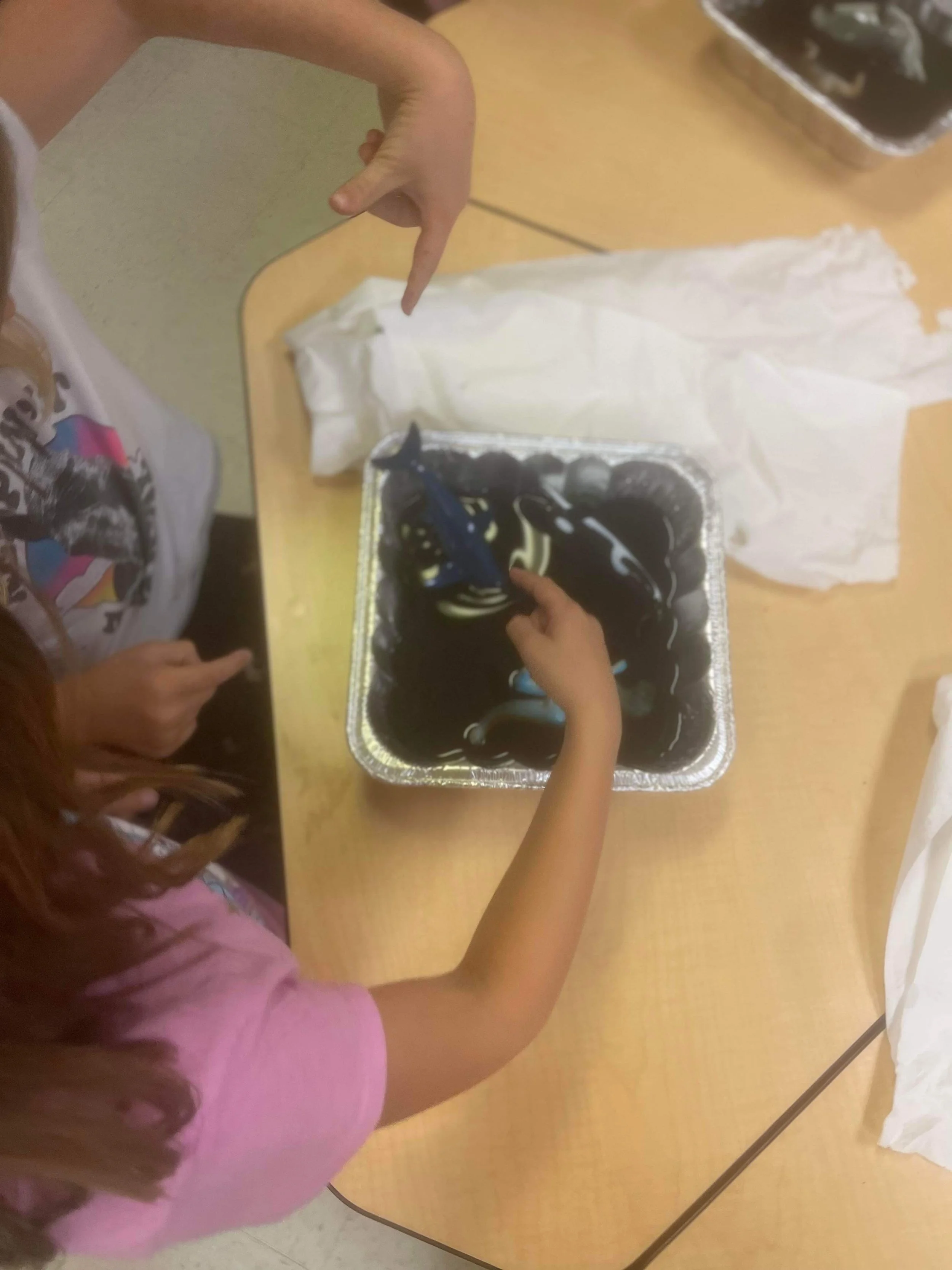Children's hands stirring black and white marbled slime in a aluminum tray on a table.