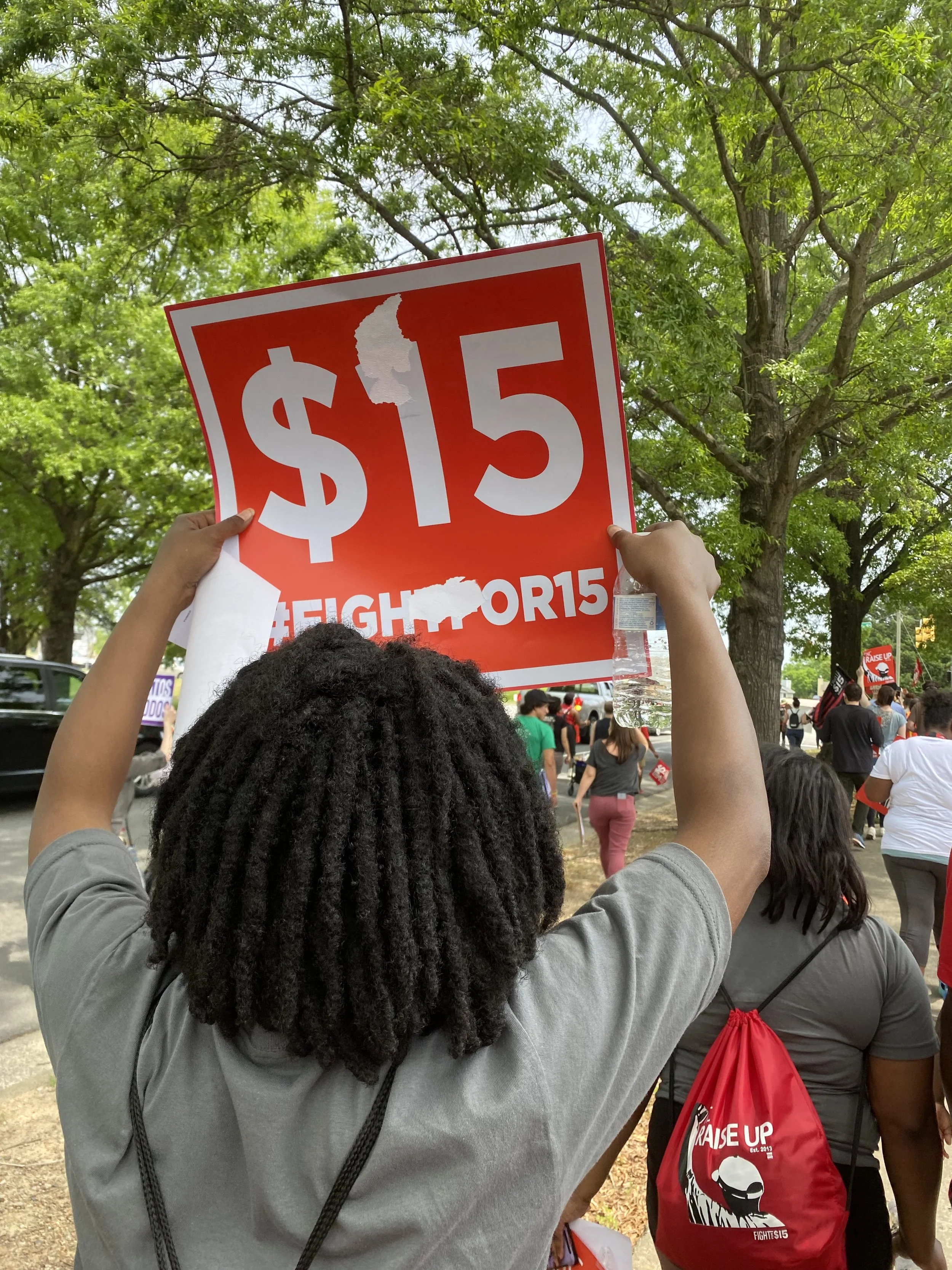 Person holding a red sign with white text displaying '$15' during a protest or rally, with other protesters walking in the background, some carrying backpacks and signs, and surrounded by trees.