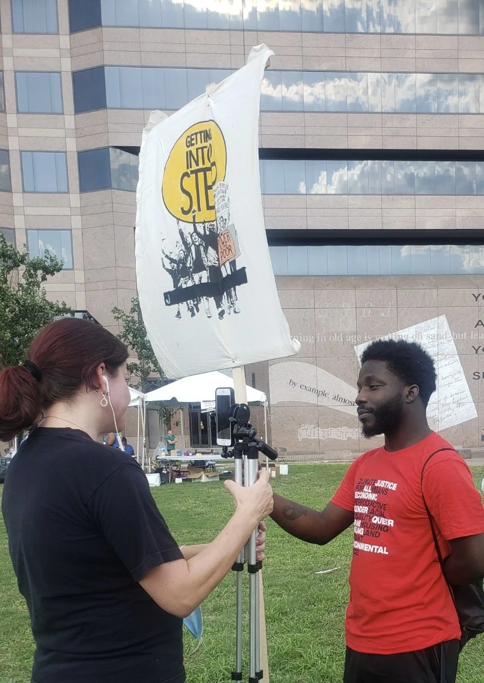 A woman and a man are standing outdoors on grass with a building in the background. The woman is holding a camera or phone on a tripod, and the man is holding a flag with the text 'Getting Into STIP' and protest imagery. The woman has earphones, and