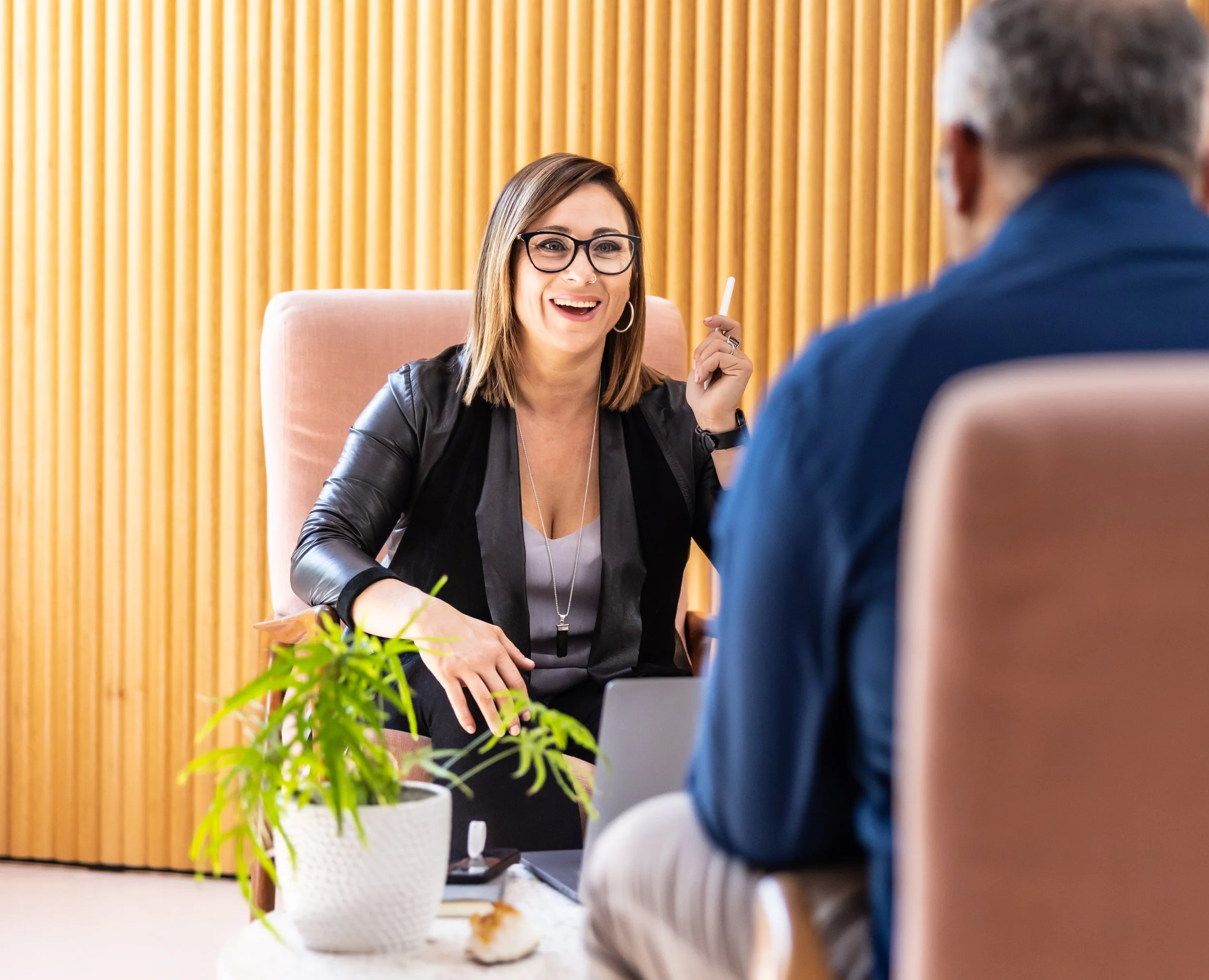 Sarah with glasses, wearing a black leather jacket, sitting on a pink chair, talking to a man with gray hair, wearing a blue blazer, in a modern office setting with wooden paneling background.