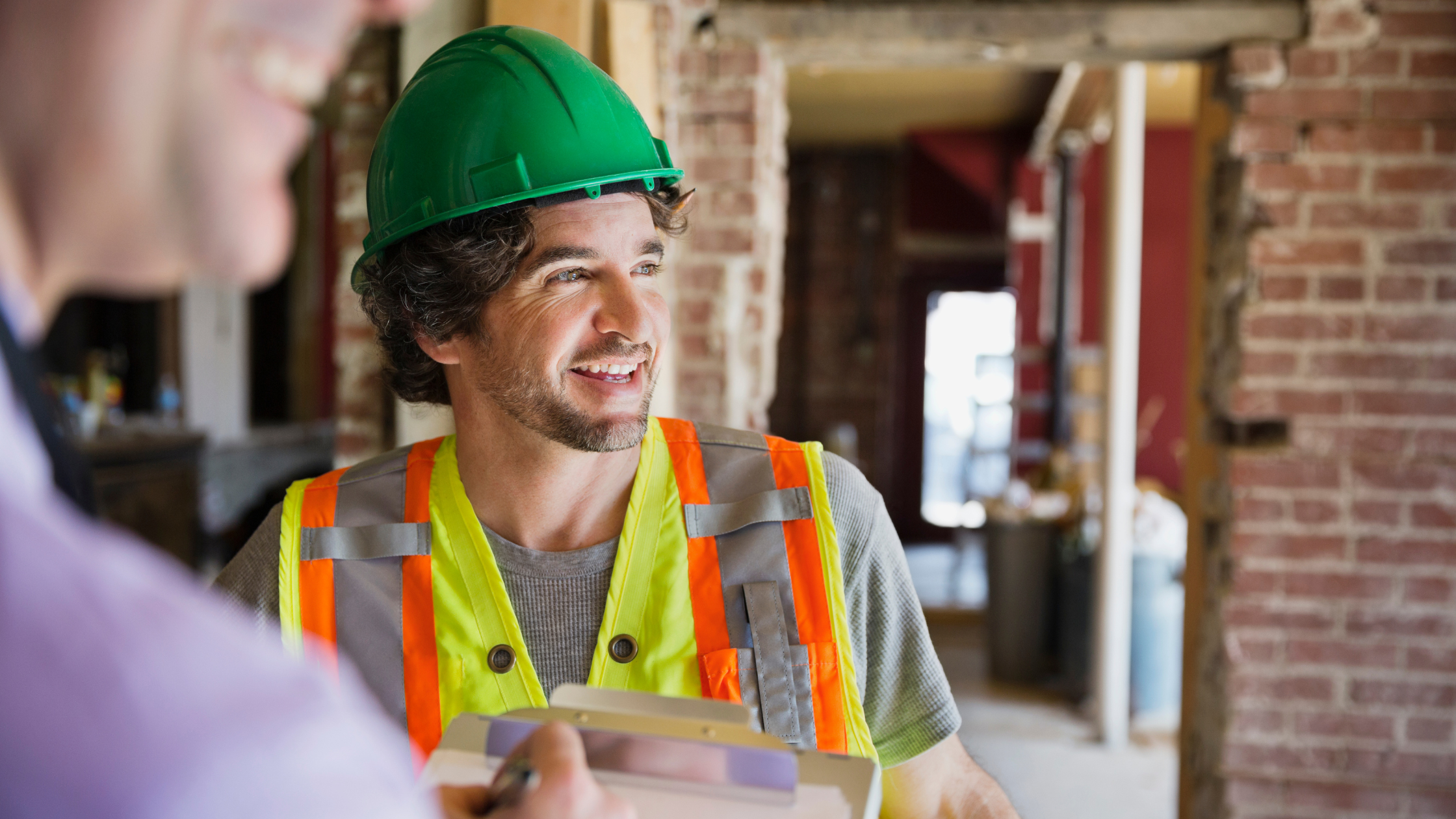 A construction worker wearing a green hard hat and a reflective safety vest smiling and talking to a person with a notepad, inside a building under construction with brick walls.