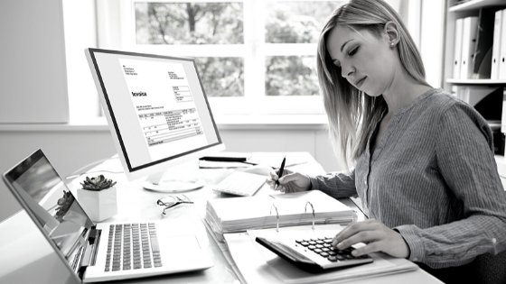 A woman working at her desk with a computer, makeup, and a calculator, in a bright office with large windows.