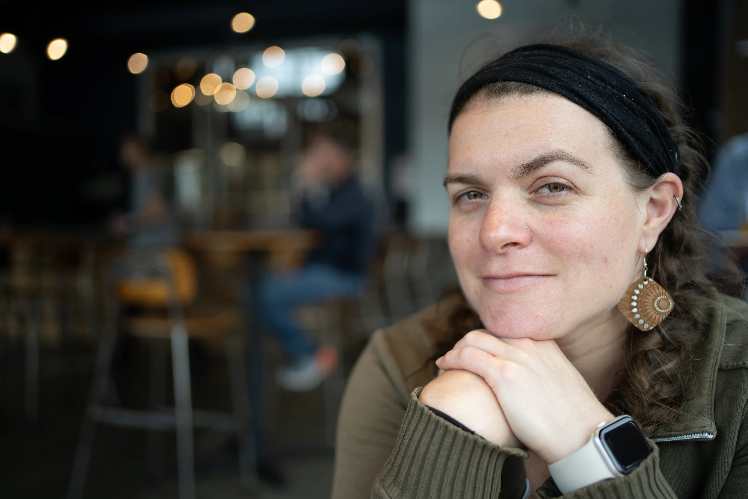 Woman with a headband smiling indoors, wearing a smartwatch and wooden earrings, with blurred background of a café setting.
