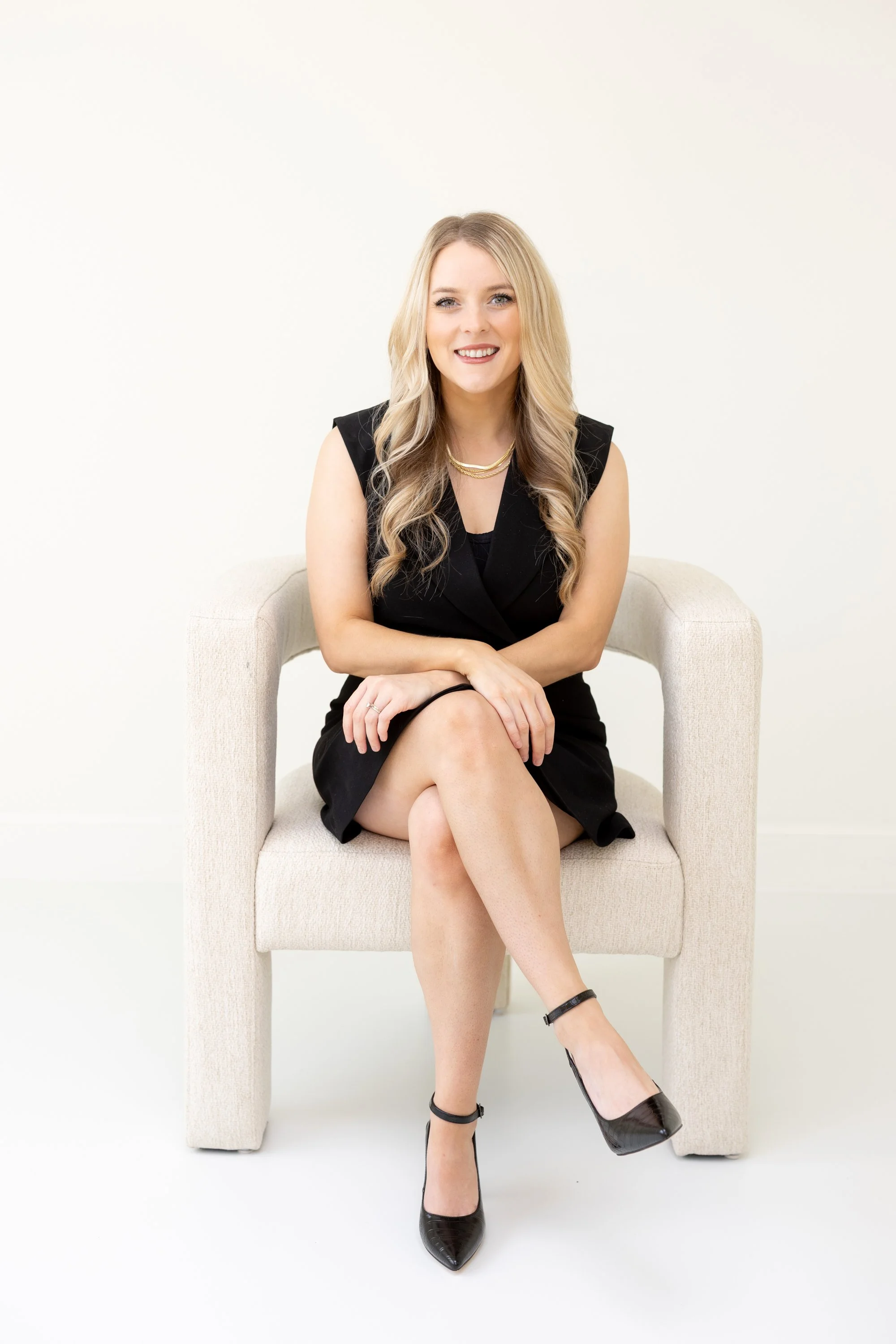A woman sitting on a beige armchair against a white background, wearing a black sleeveless dress, black high-heeled shoes, and gold jewelry, smiling at the camera.