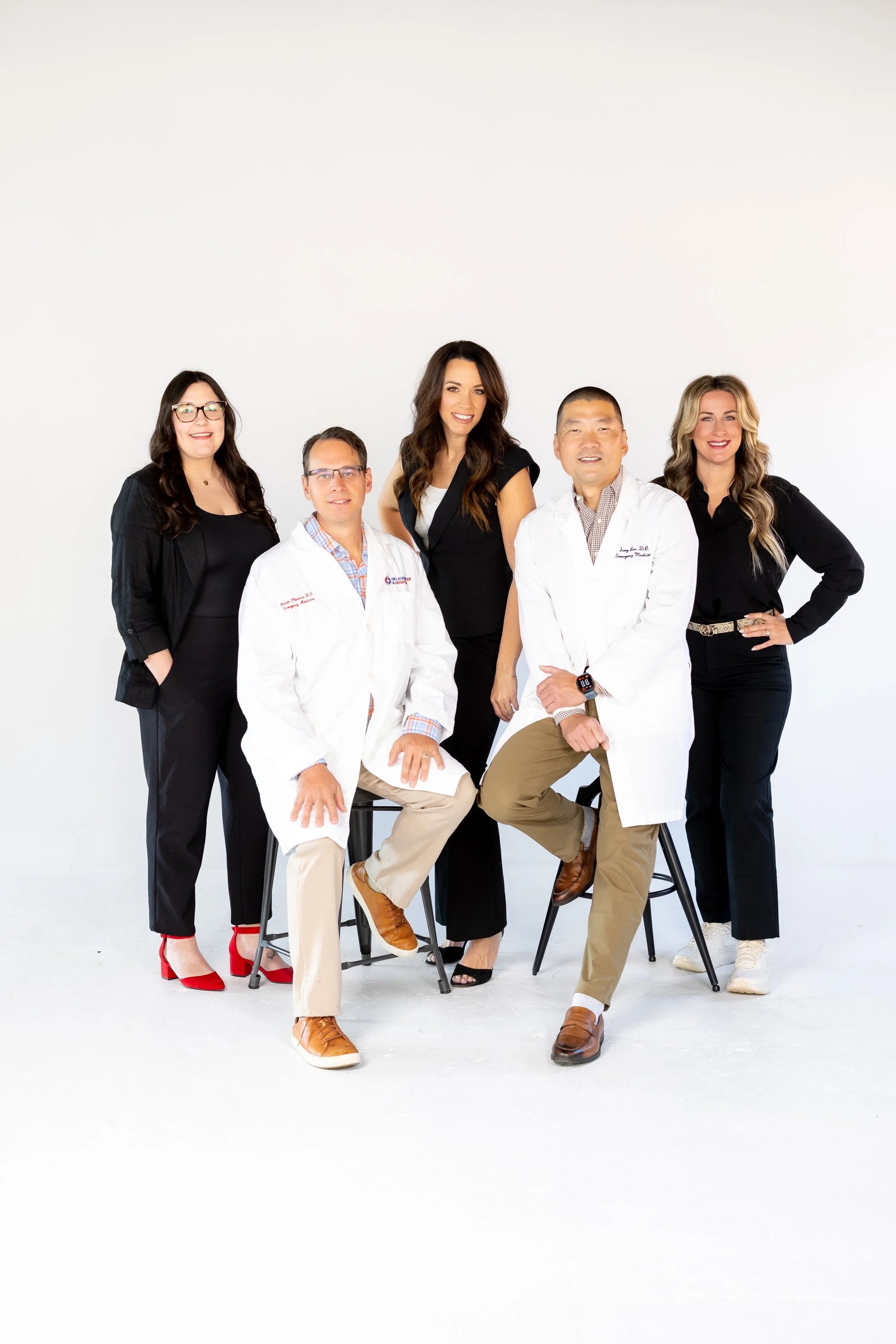 Group of six diverse professionals, including two seated in front and four standing behind, posing against a white background. They are dressed in business casual attire, with some wearing white coats, indicating a medical or healthcare setting.
