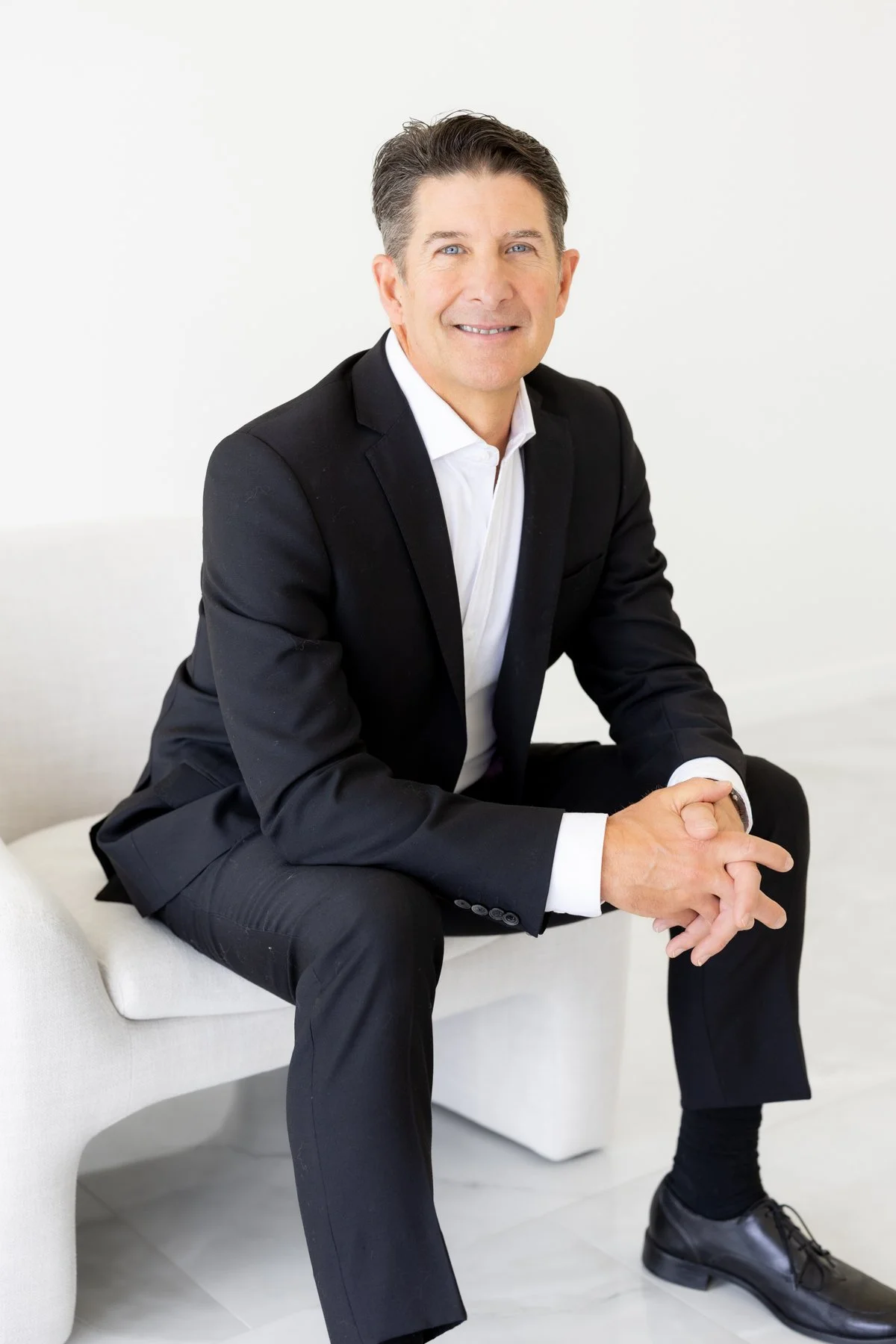 A man in a black suit sitting on a white couch, smiling, with hands clasped together.