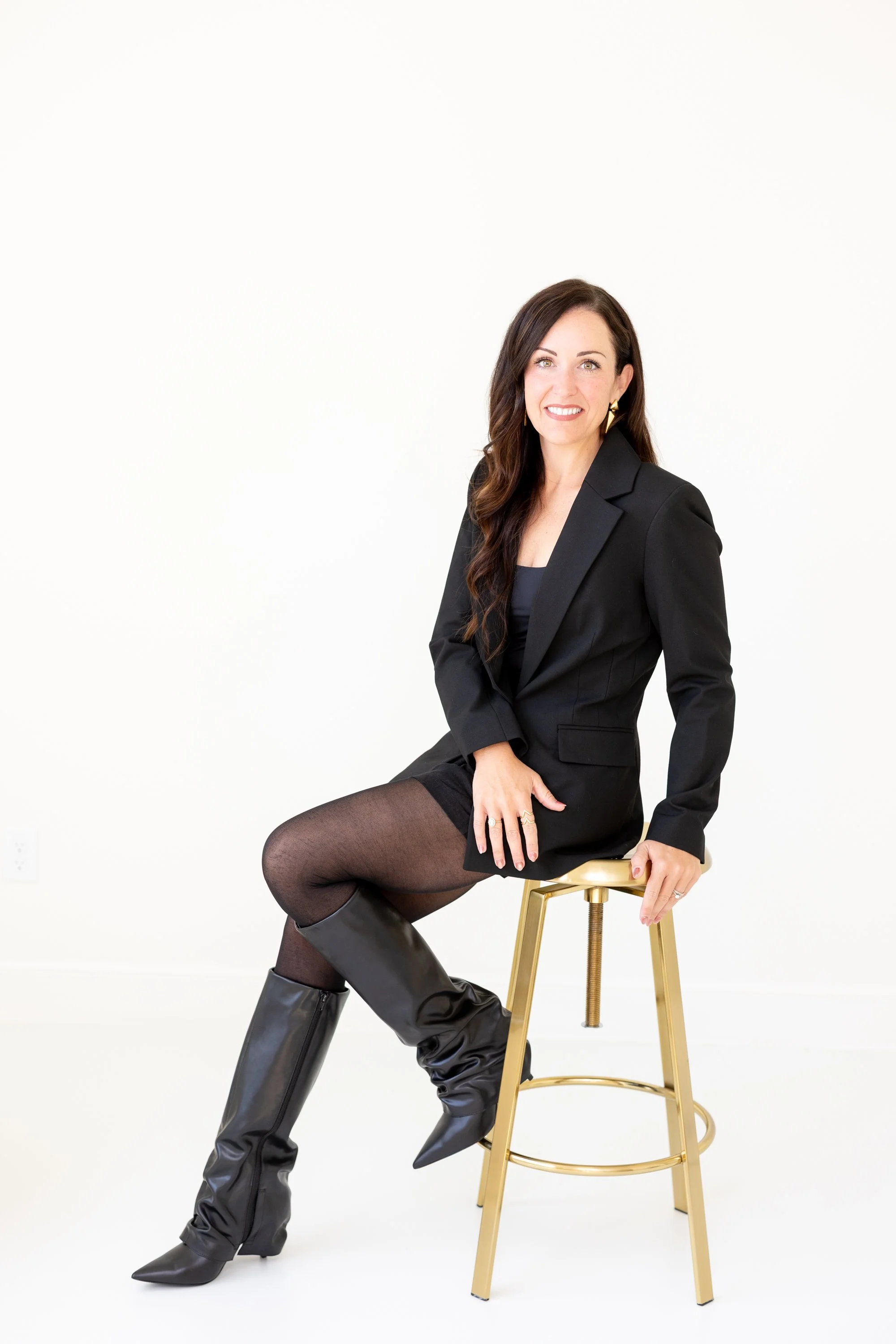 A woman with long dark hair, wearing a black blazer, black skirt, black tights, and black knee-high boots sitting on a gold stool against a plain white background.