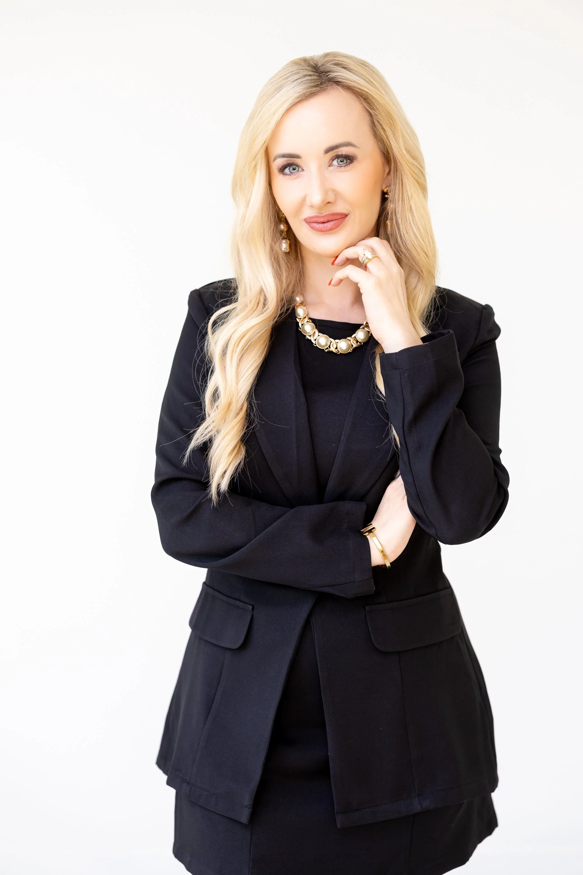 Woman in black blazer and skirt, wearing gold jewelry, with blonde hair, posing against a white background.