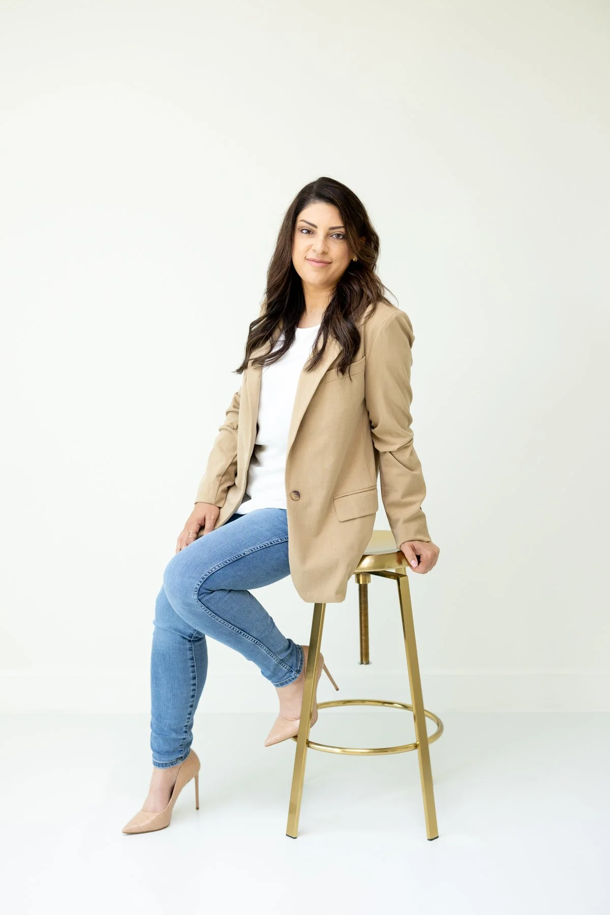 A woman with dark hair wearing a beige blazer, white top, and blue jeans, sitting on a gold stool with one leg crossed over the other, smiling at the camera.
