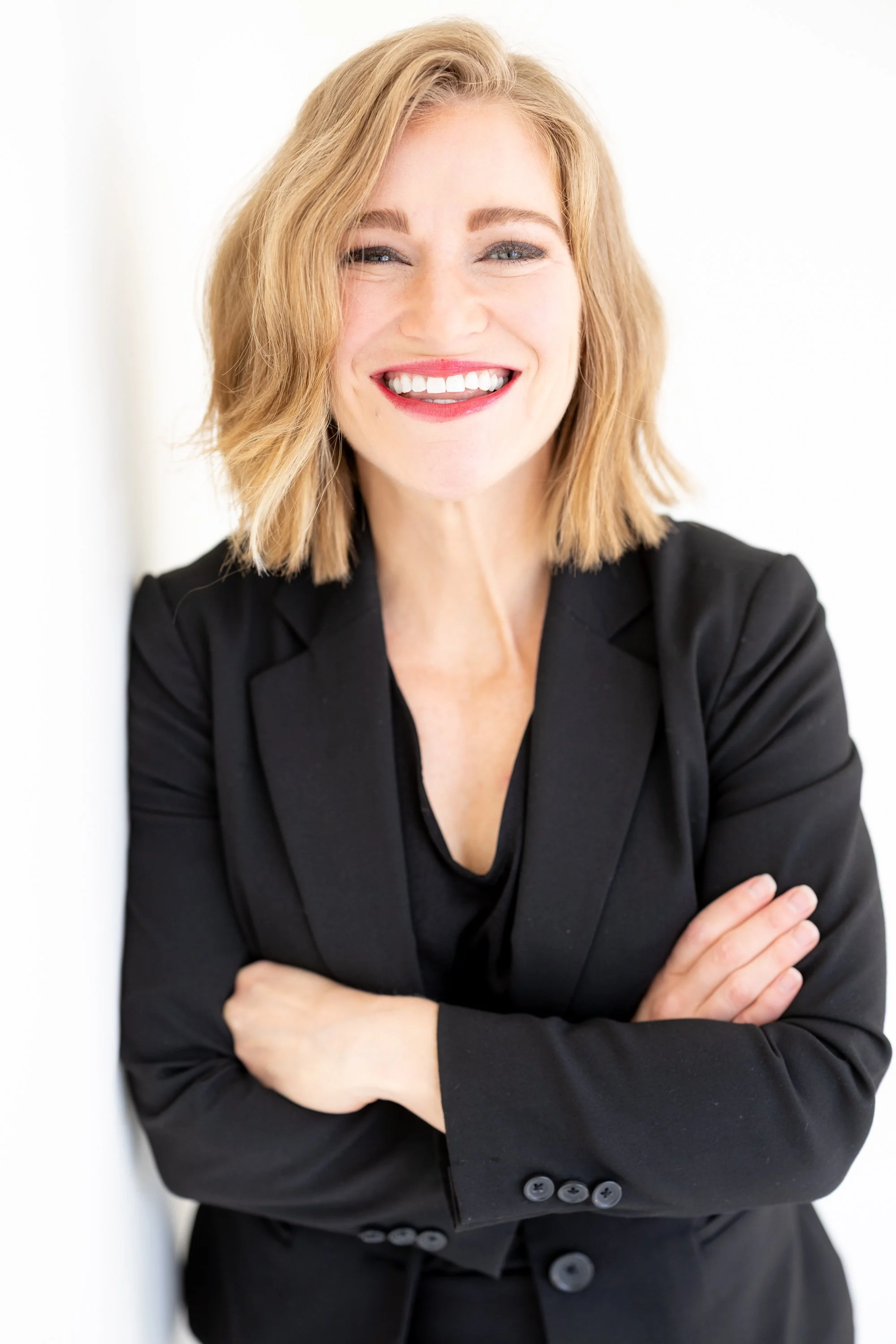 A confident woman smiling with arms crossed, wearing a black blazer, standing against a white background.