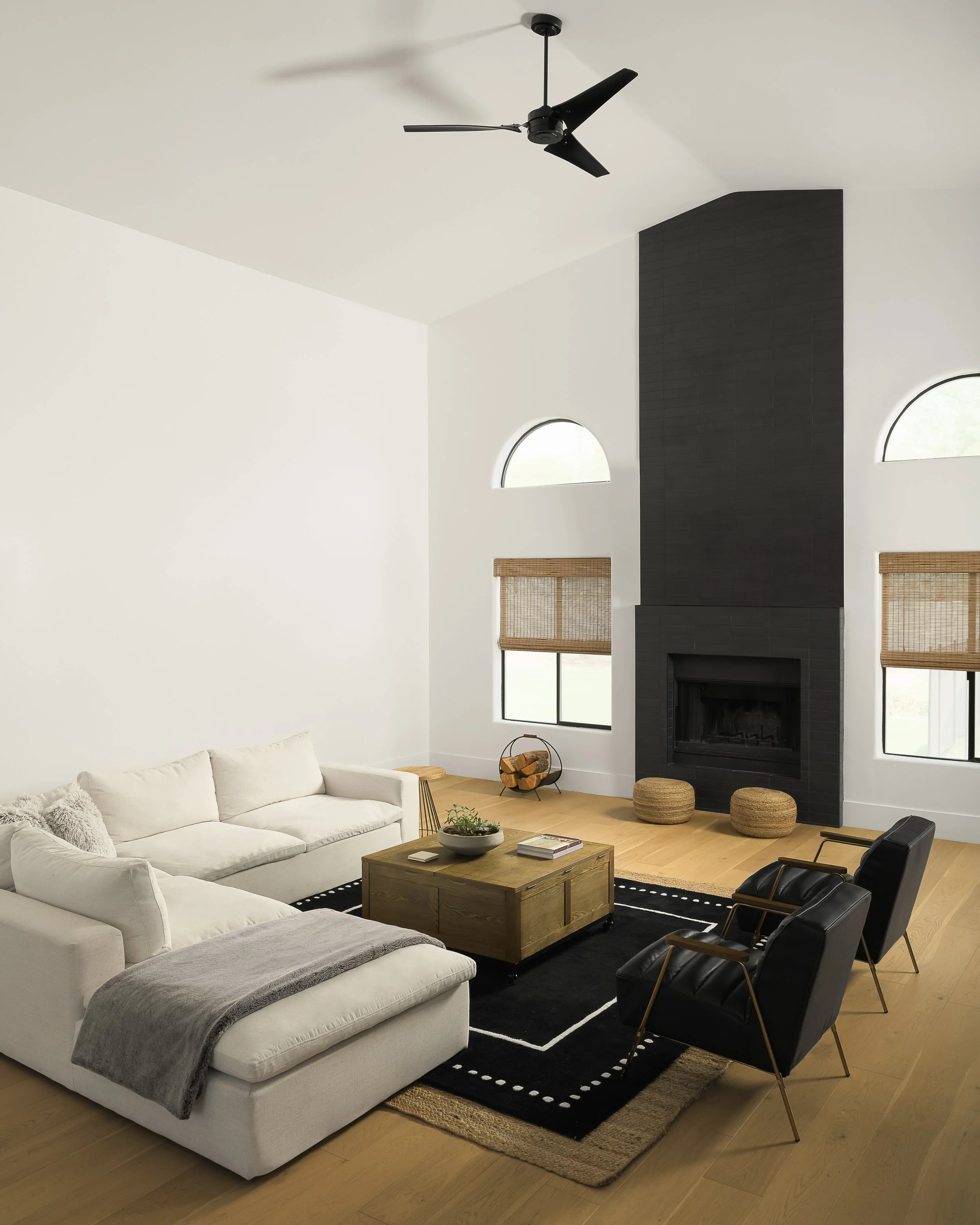 Living room with white sectional sofa, black and wooden chairs, black fireplace, natural wood coffee table, and large windows with blinds, ceiling fan overhead.