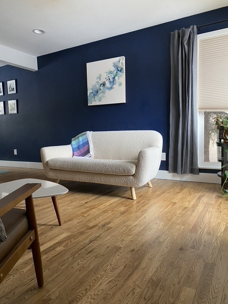 Living room with a cream-colored couch, a colorful striped blanket, blue wall with artwork, window with curtain, and wooden flooring.