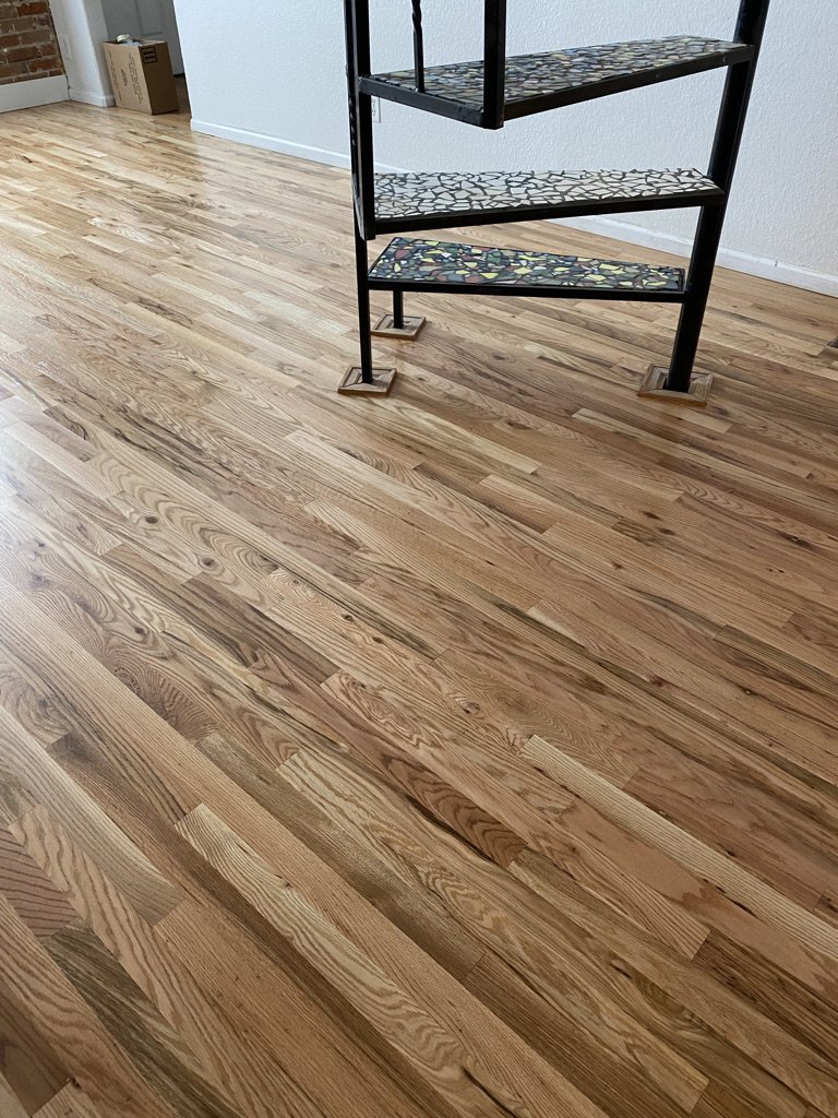 Photo of a hardwood floor with a black metal shelving unit with mosaic tile shelves in a room with white walls and a cardboard box in the corner.