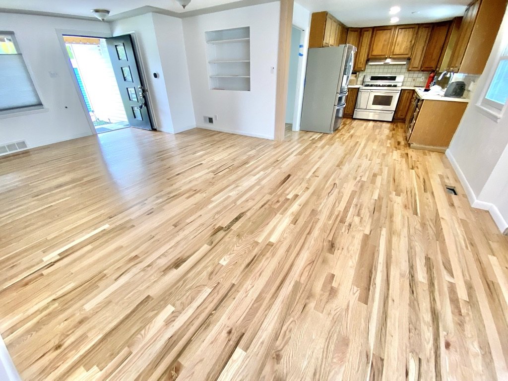 Empty living room with hardwood floors and open kitchen with wooden cabinets and appliances.