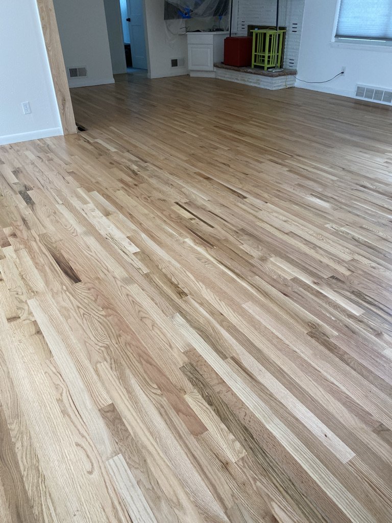 Smooth, polished hardwood floor in a room with white walls and a window, with some furniture and household items in the background.