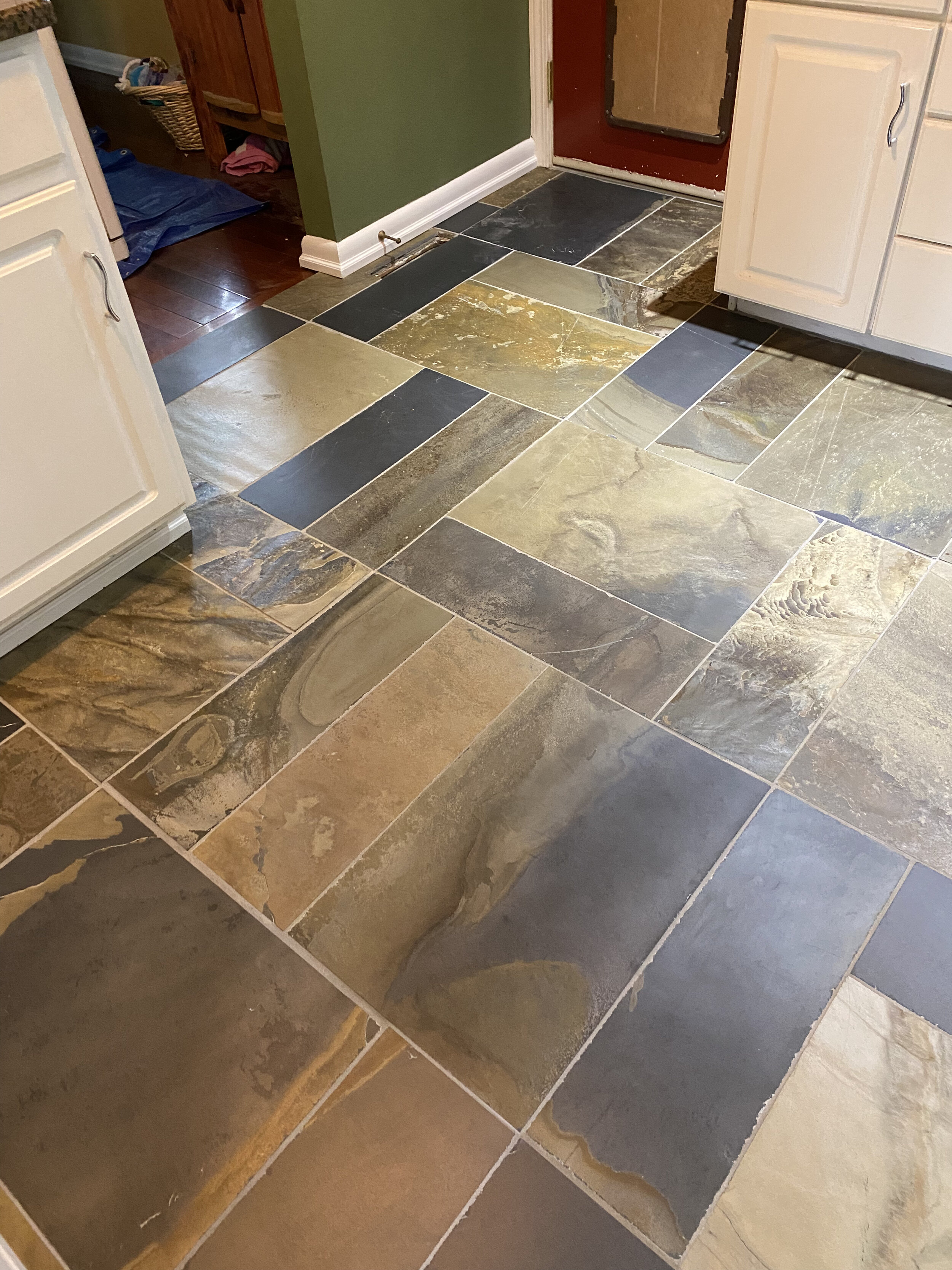 A kitchen floor with multicolored slate tiles in shades of brown, gray, black, and tan. The tiles are arranged in a staggered pattern around a doorway and cabinets.