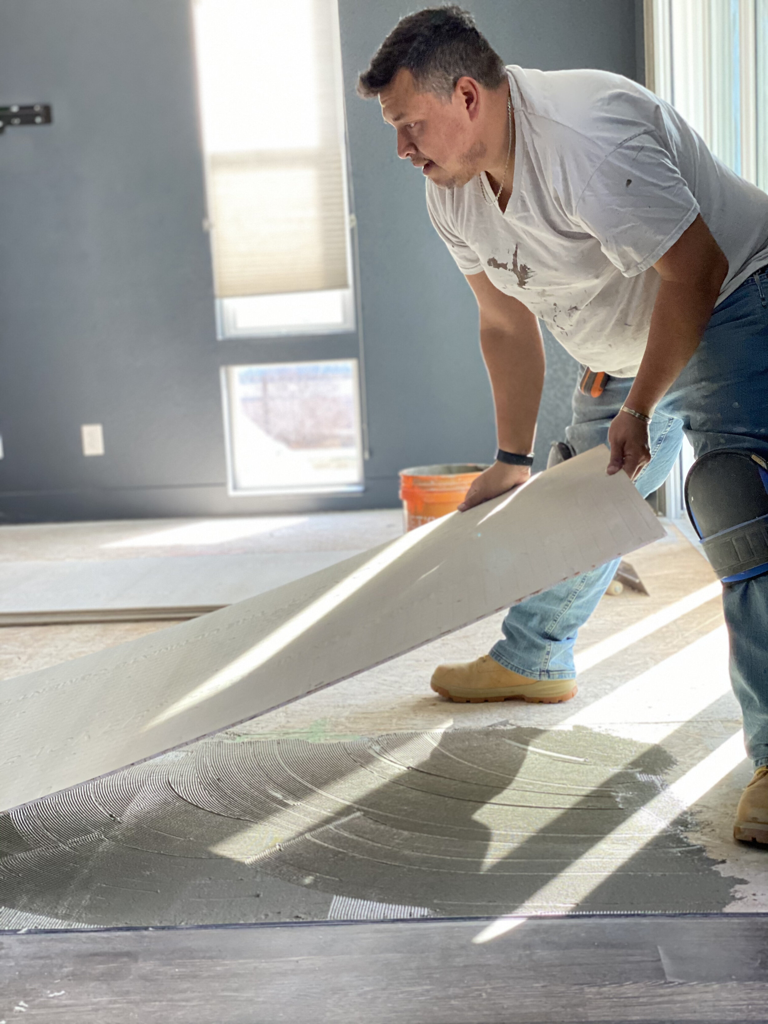 A man installing flooring, holding a piece of flooring material, on a subfloor with a foam underlayment, in a room with blue walls and large windows.