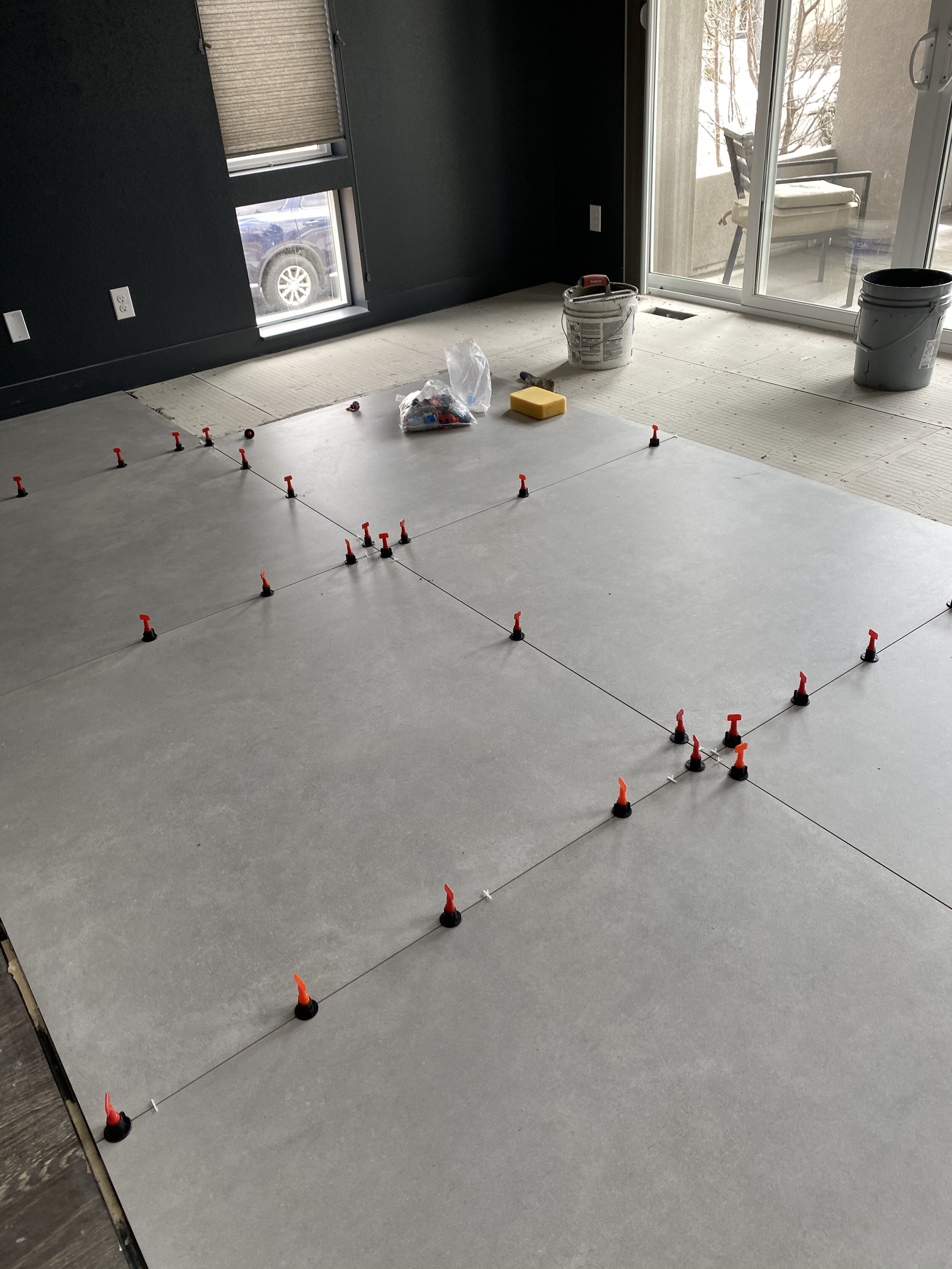 Room under renovation with tile flooring being installed, visible spacers, tools, and supplies scattered around.