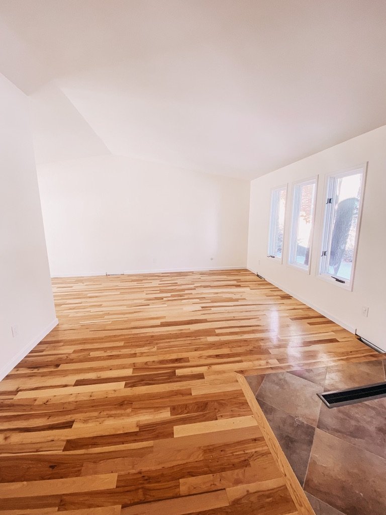 Empty living room with white walls, hardwood floors, and three large windows showing outdoor trees; corner with tiled area and vent.
