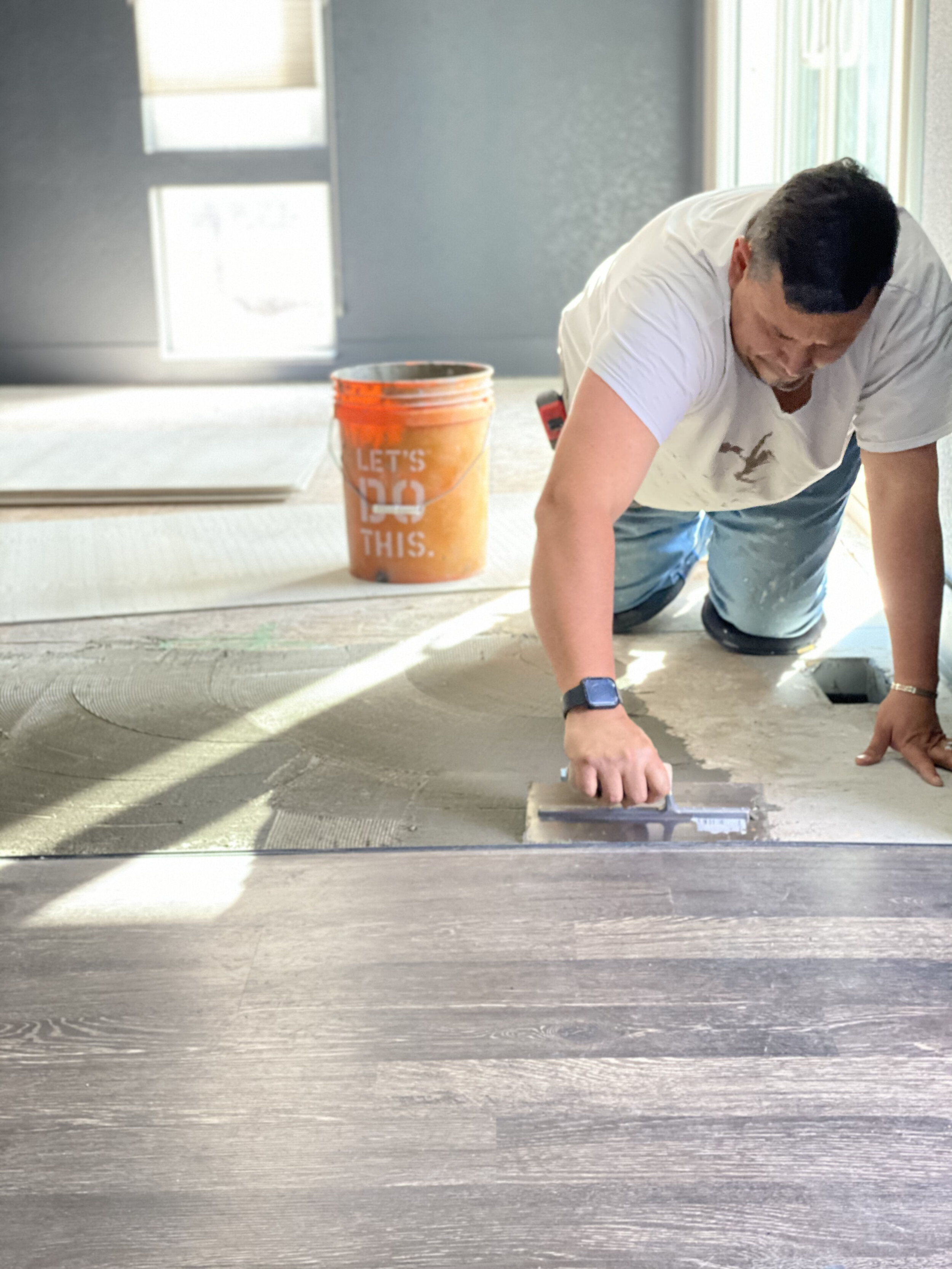 A man installing or repairing a hardwood floor, kneeling on the floor, using a trowel to spread adhesive or grout, with a bucket nearby.