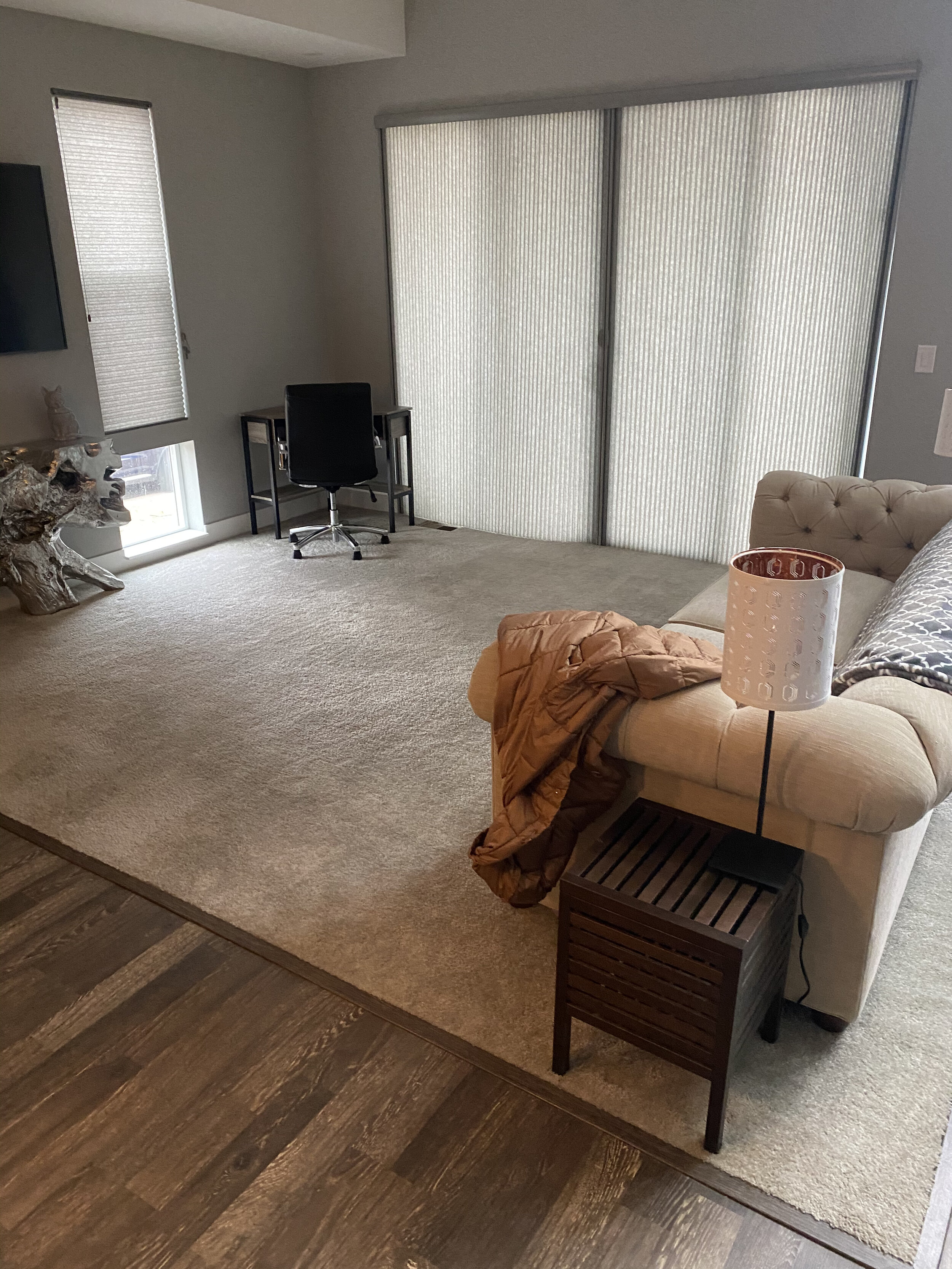 Living room with beige carpet, a beige couch with a brown jacket on it, a side table with a lamp, a small black desk with a black chair, large sliding glass doors with vertical blinds, a small window with a vertical blind, and a mounted flat-screen T
