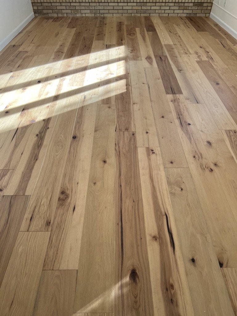 Wooden floor with sunlight and shadow patterns, with a brick wall and white walls in the background.