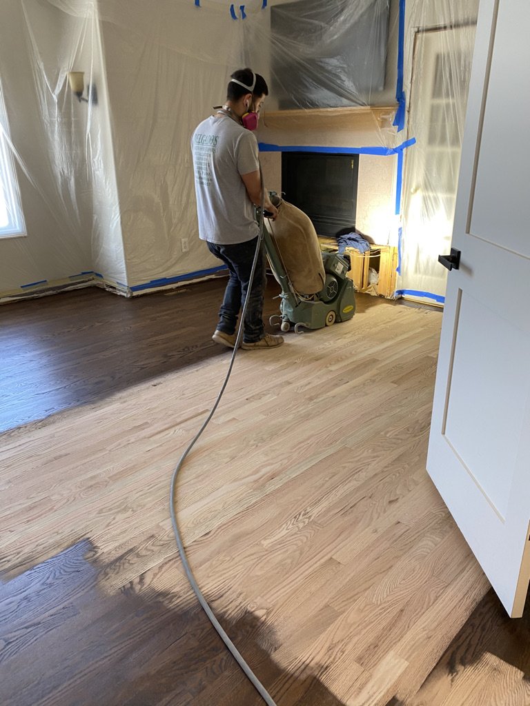 Person using floor sanding equipment in a room with partially sanded wooden floor, plastic sheeting and painter's tape covering walls and fireplace.