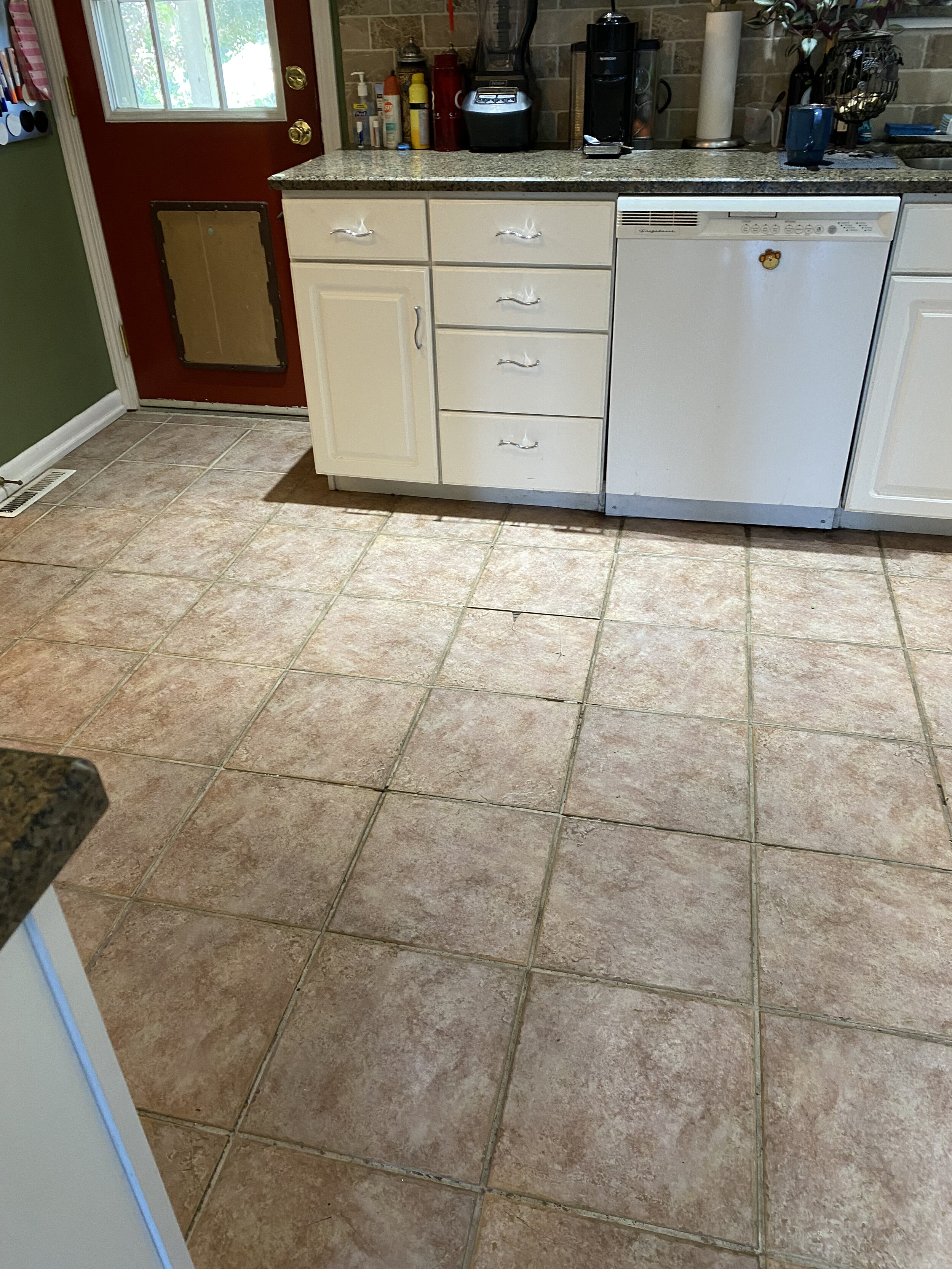 Kitchen floor with pinkish-brown tiles, white cabinets, countertop with various appliances, and a red door with a pet door installed.