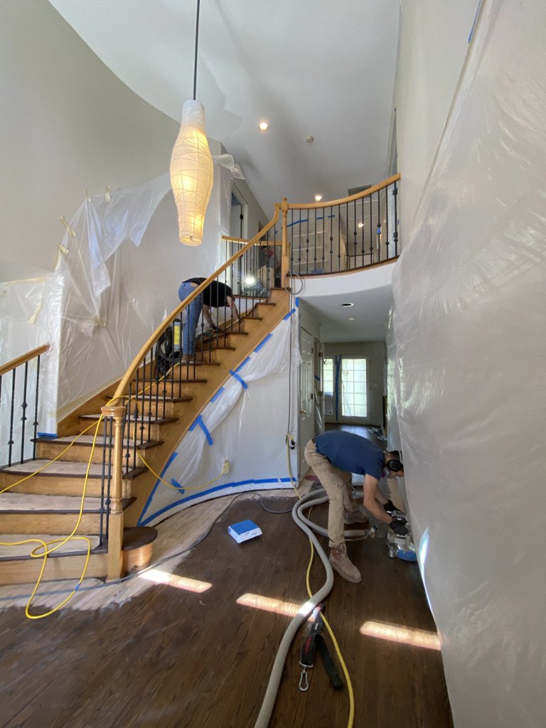 Interior of a house under renovation with plastic sheeting taped along the curved staircase and walls, a worker on the floor using tools, and equipment including hoses and extension cords.