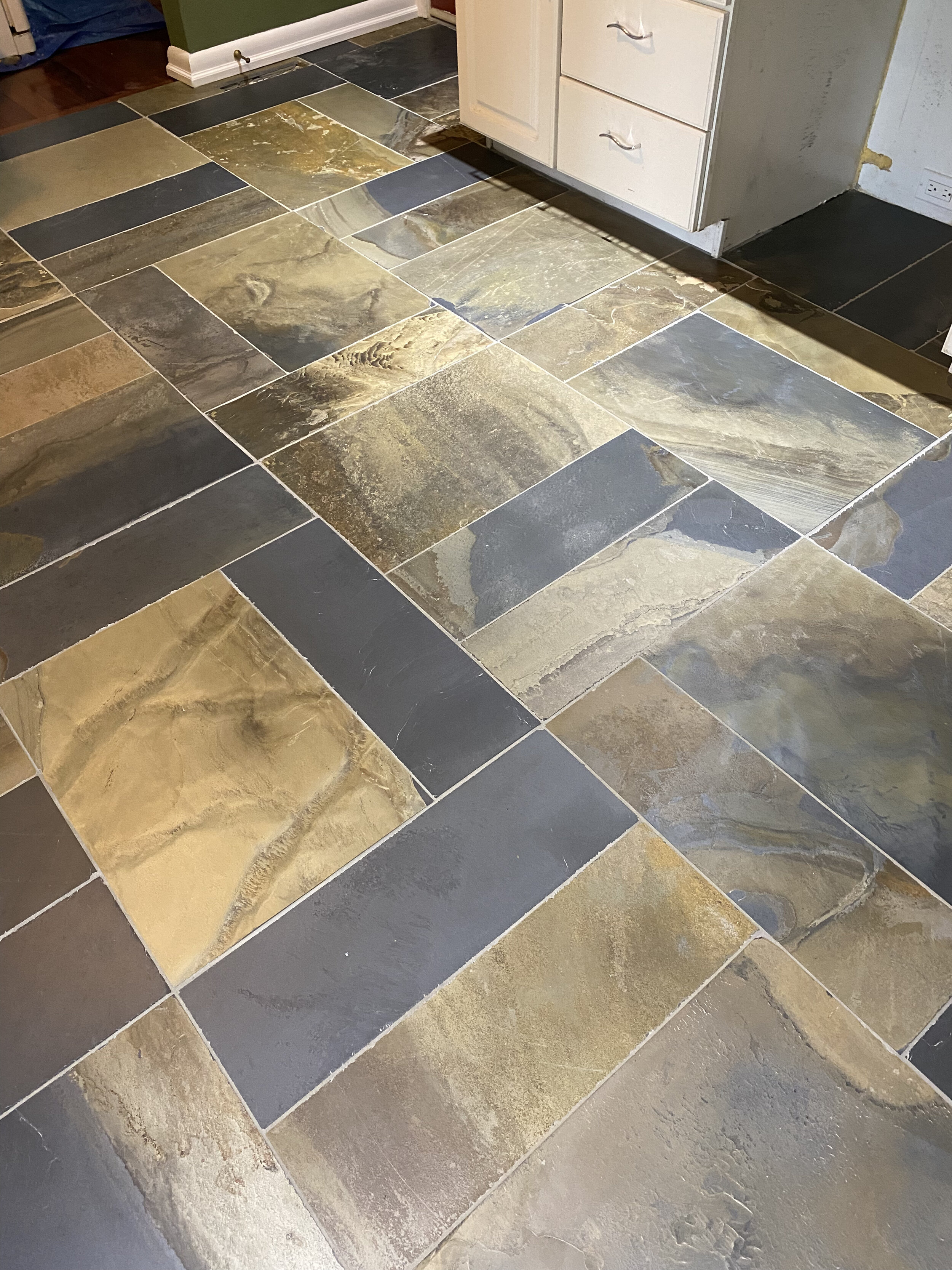 Colorful natural stone tiles arranged in a geometric pattern on a kitchen floor, next to white cabinets and a green wall.