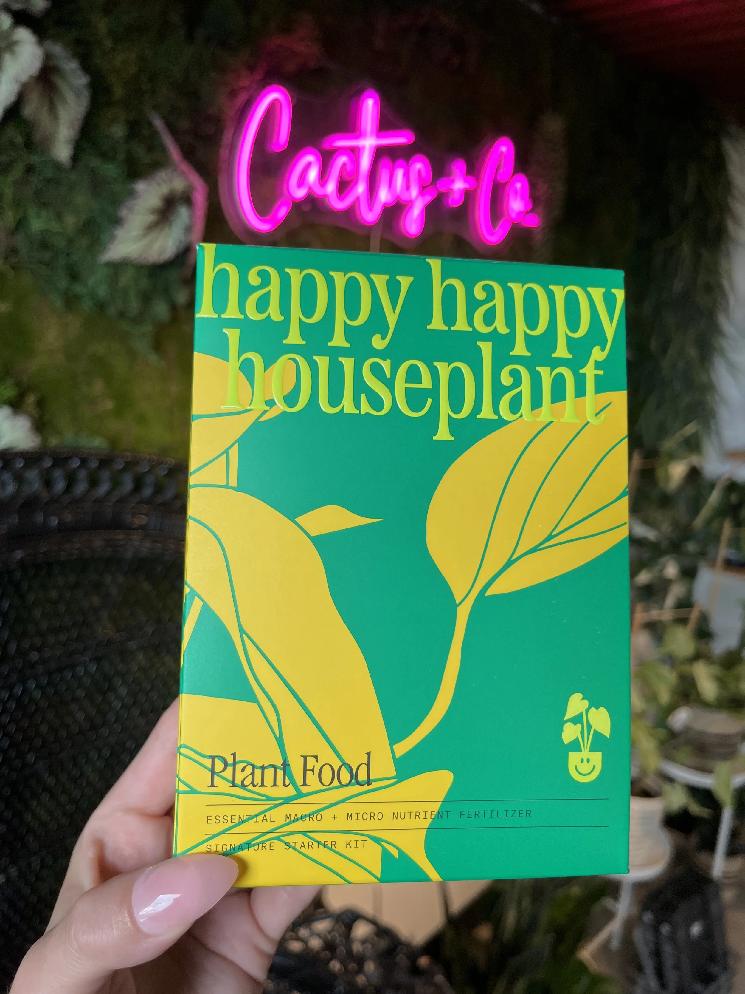 A hand holding a colorful box of 'happy happy houseplant' Plant Food  in front of a wall with a neon pink sign reading 'Cactus + Co'
