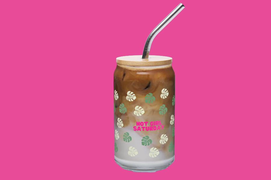 A glass jar with a lid and a metal straw contains an iced coffee beverage with milk, with pink text that says "Hot Girl Saturday" and a pattern of green and white monstera leaves, set against a pink background.