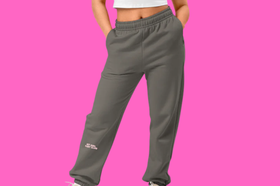 Person wearing gray sweatpants that say "Hot Girl Plant Club' with hands in pockets against a pink background.