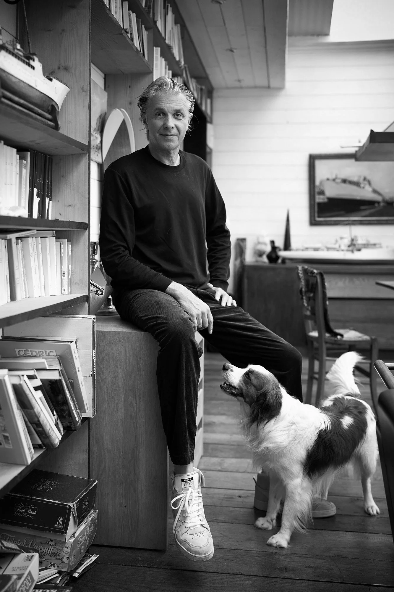 Ein Mann sitzt auf einem Bücherregal, mit einem Hund, der zu seinen Füßen steht, in einem Raum mit Bücher, Möbeln und Wandkunst.