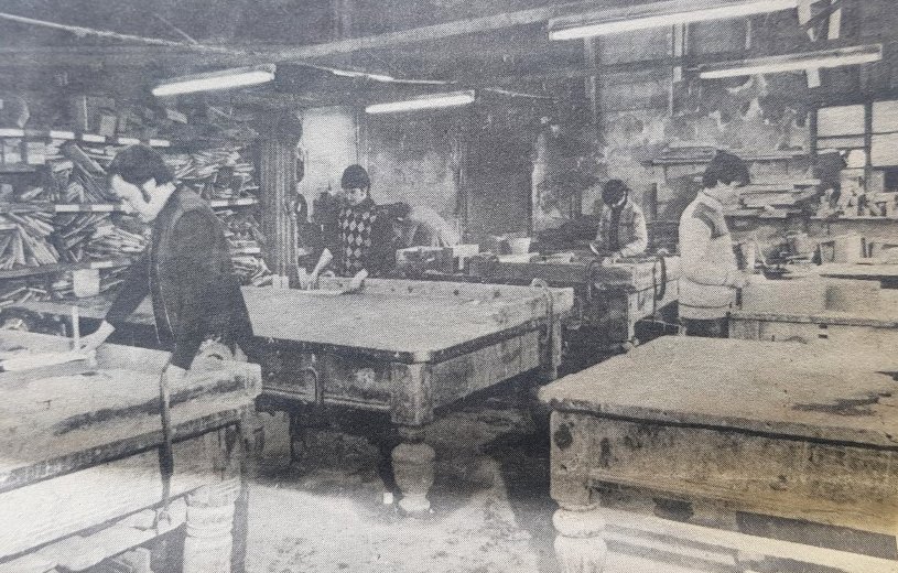 Vintage black and white photograph of people working in a manual labor workshop, with wooden tables and materials around the room.