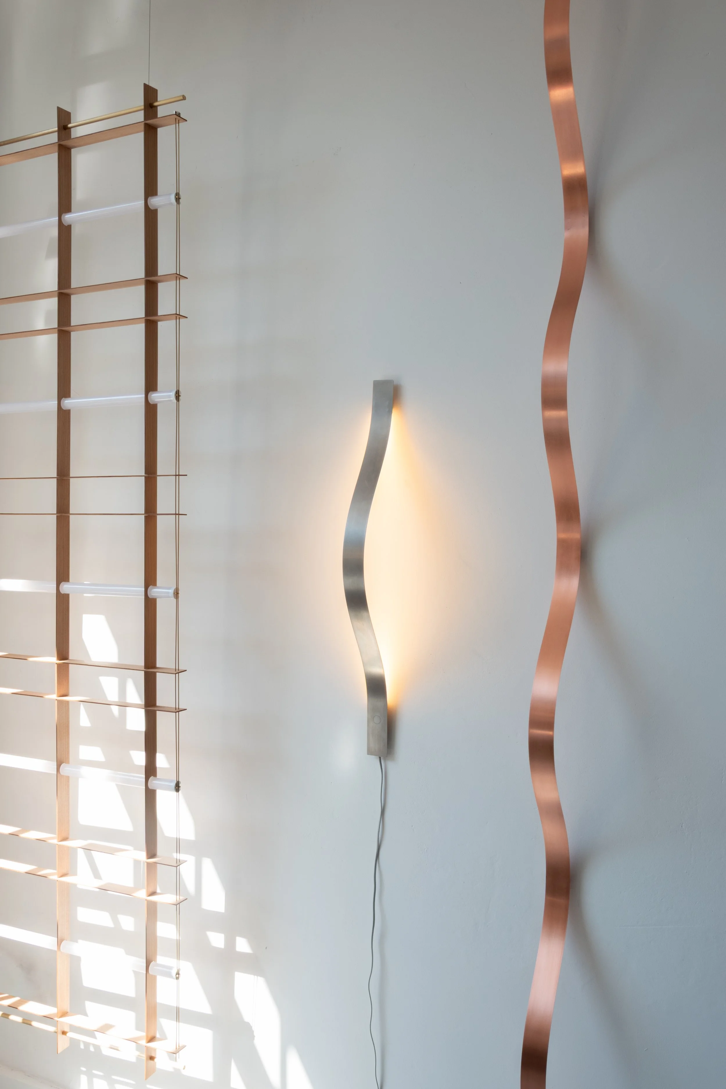 Modern wall decor featuring a wooden grid structure with horizontal slats on the left, a wavy metallic sculpture in silver in the center, and a tall, slender curved metallic sculpture in copper on the right, all mounted on a plain light-colored wall with soft lighting.