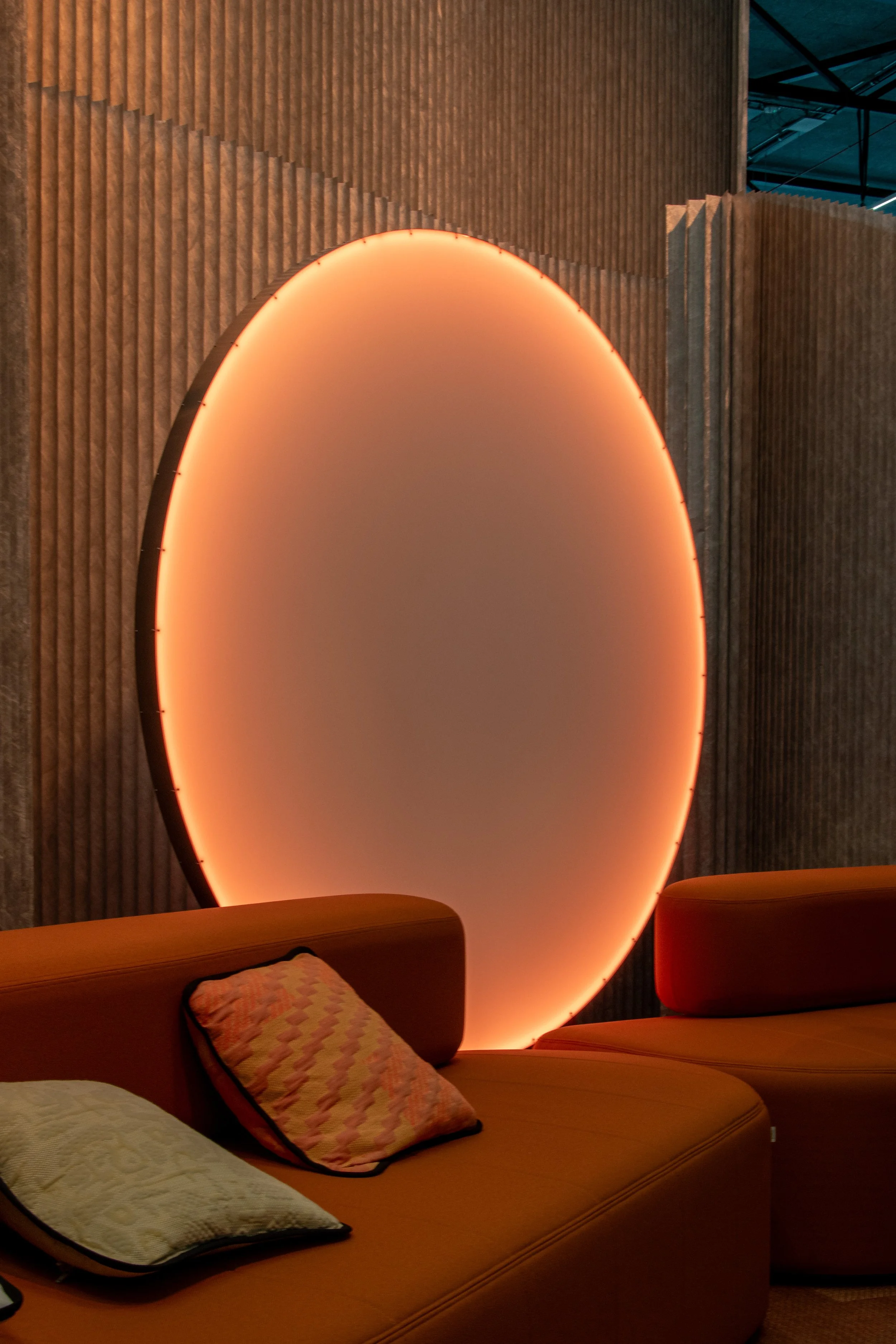 Interior of a modern room with a large, circular, backlit wall decoration emitting a warm, orange glow. There are orange rounded chairs and pillows on the seats in the foreground, with a textured wall behind the decoration.