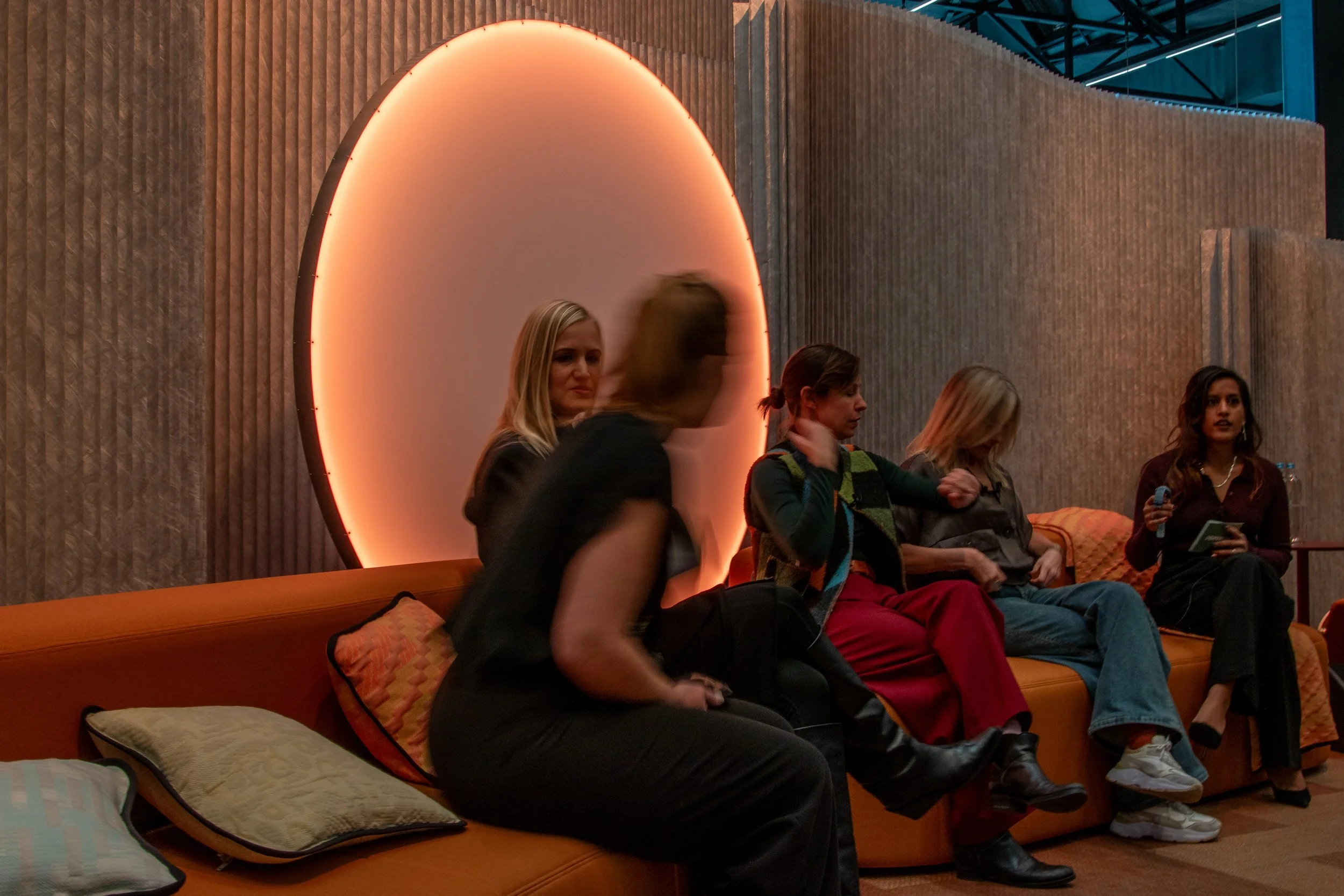 Five women sitting on an orange couch indoors, with one holding a phone and another adjusting her jacket, in front of a wall with a large illuminated circular feature.