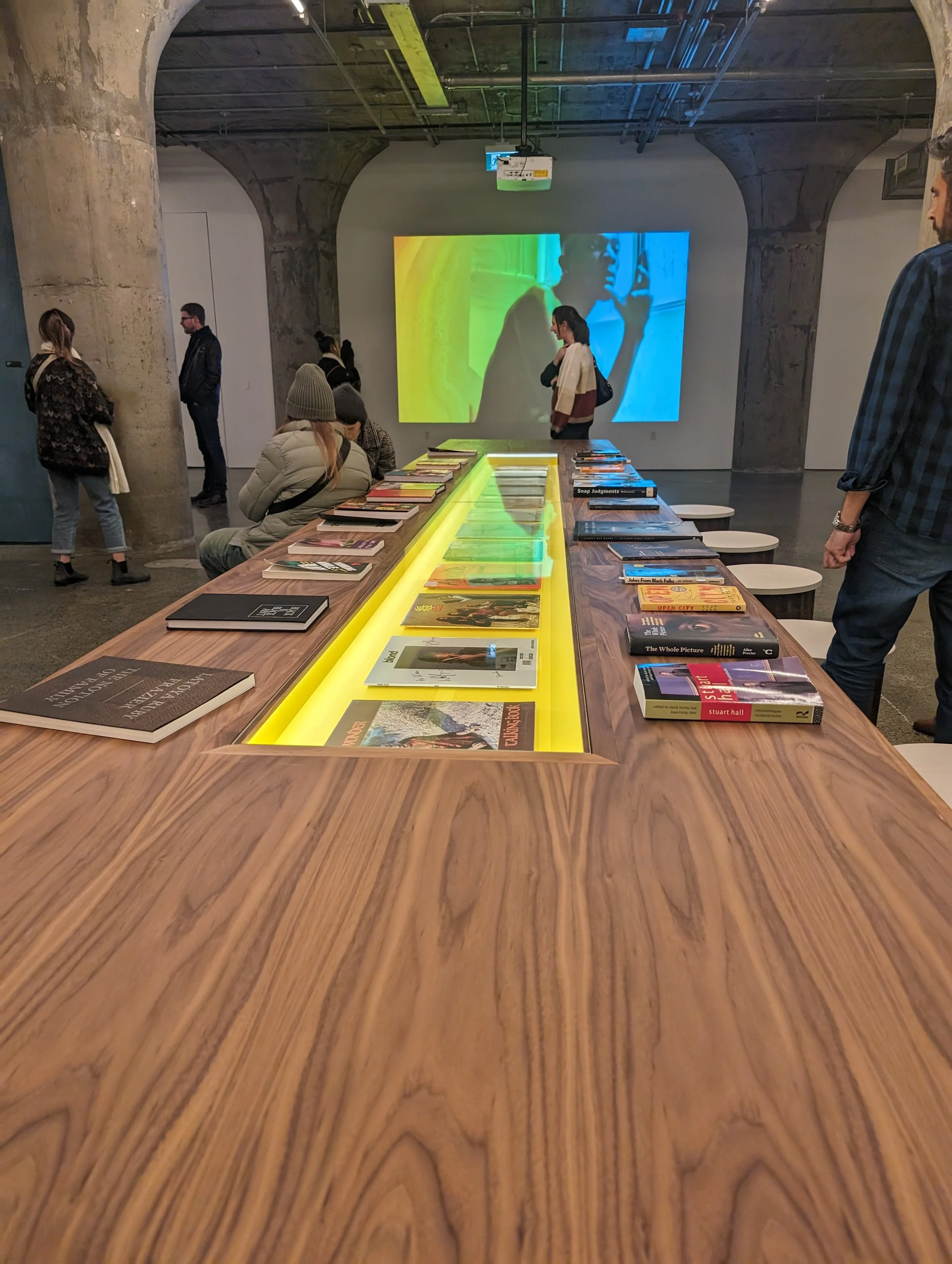 A long wooden table lined with three rows of colourful books, and the middle row accented with a yellow light. People are walking around and sitting by the table.