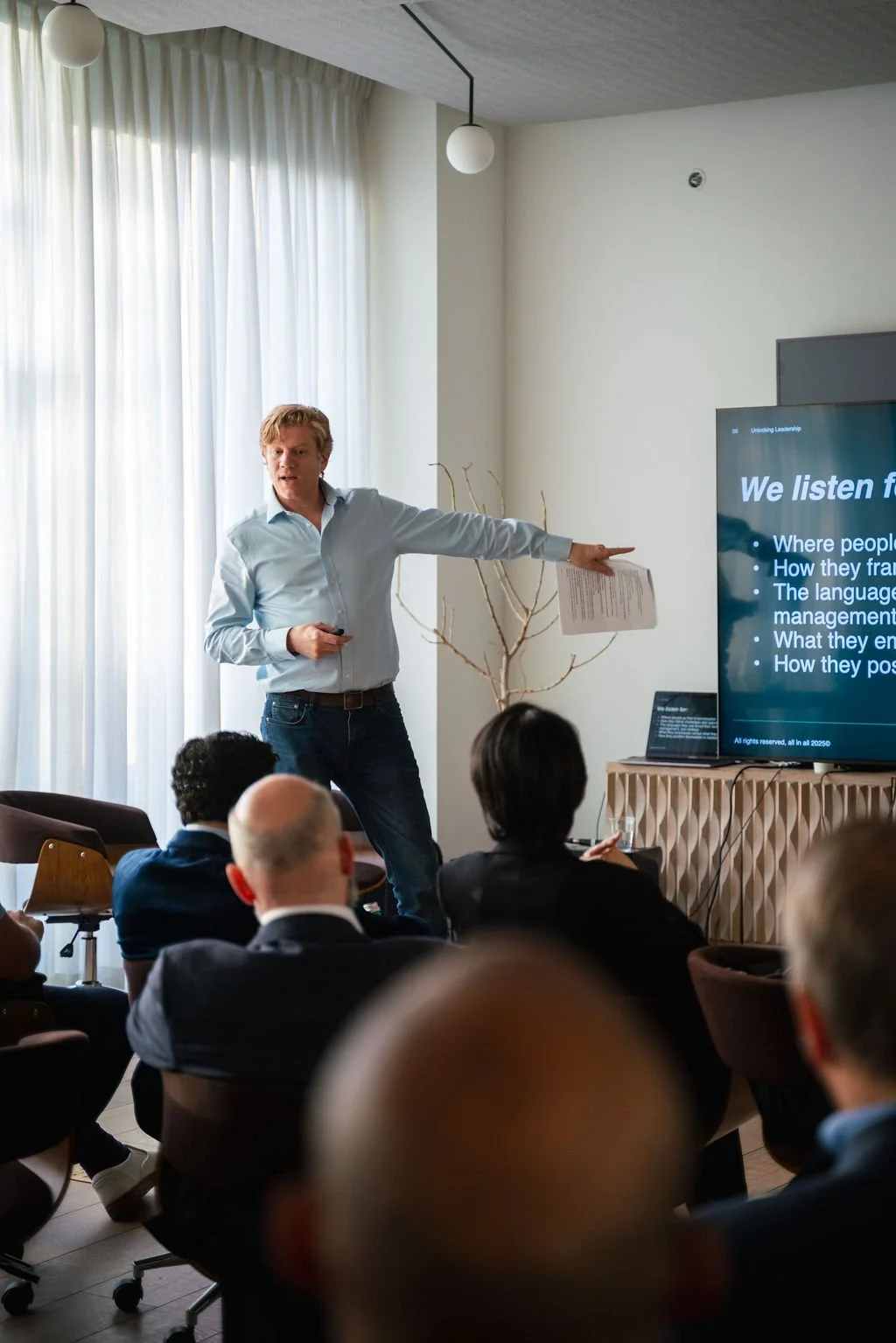 A person giving a presentation in a conference room, standing at a podium in front of a screen displaying partial text "Uncertain, Complex, Ambiguous" with an audience seated at tables.