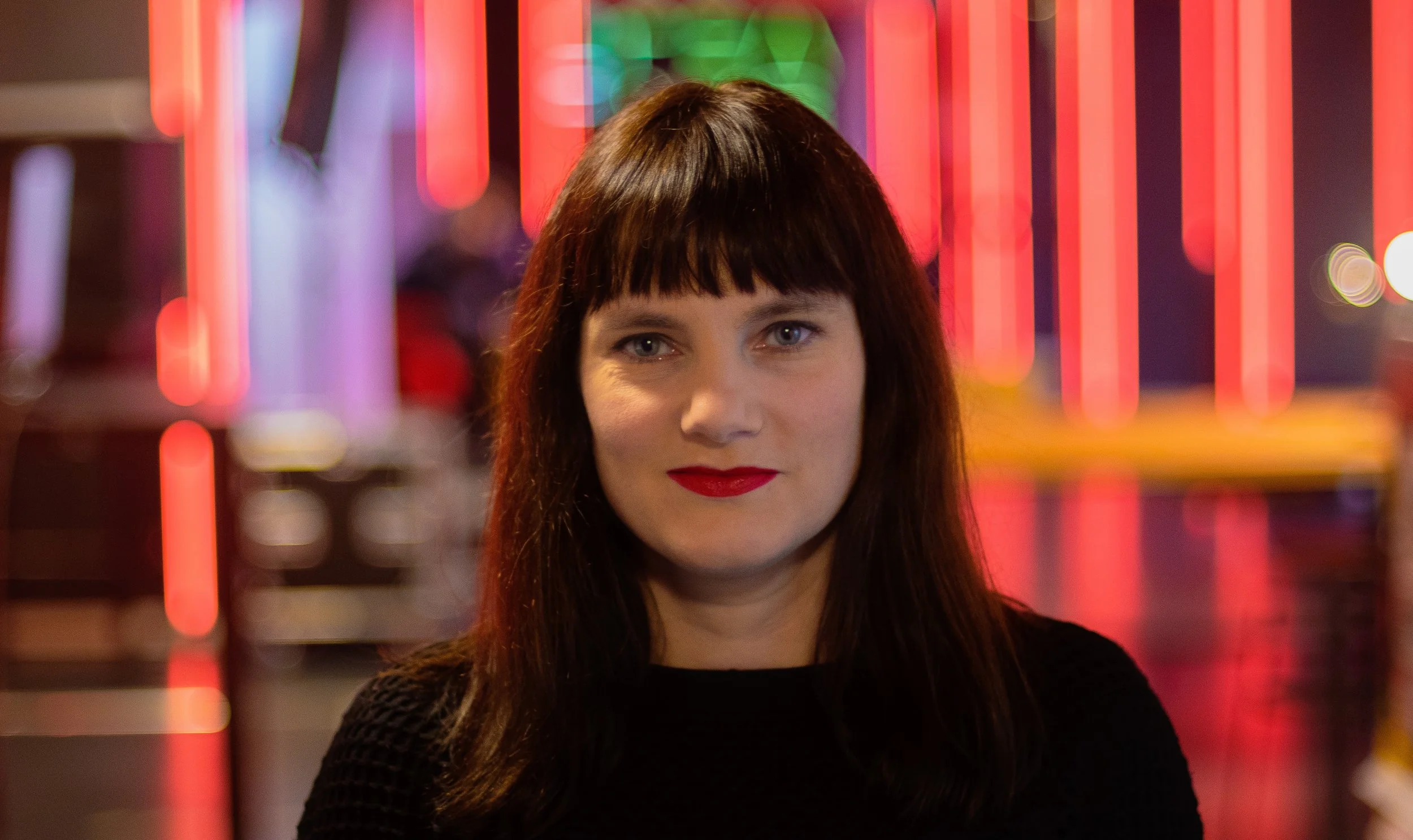 A woman musician with straight dark hair and bangs, wearing red lipstick and a dark sweater, standing in front of colourful neon lights at night.