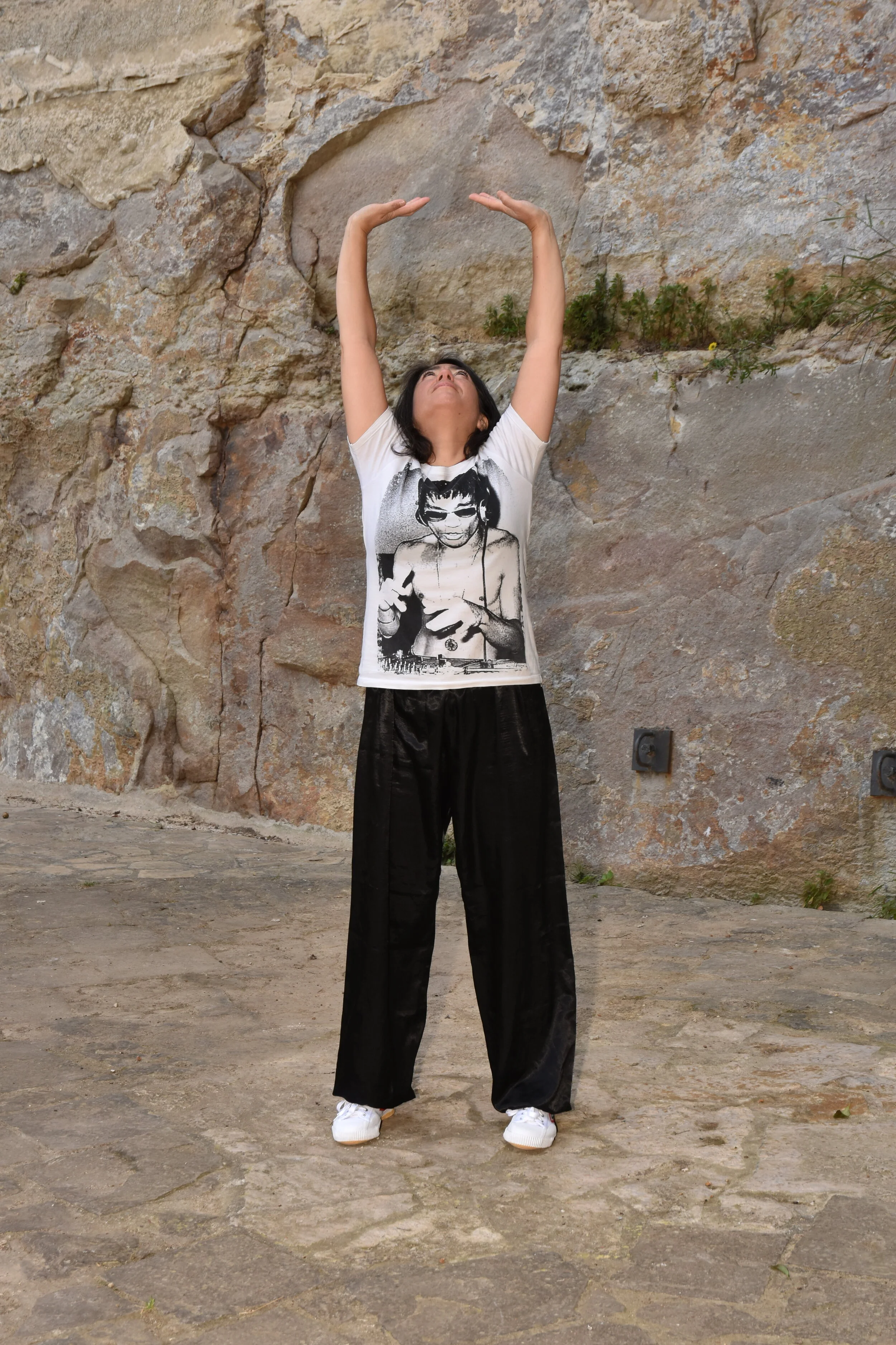 A woman standing outdoors in front of a stone wall, raising her arms above her head in a stretching pose, wearing a graphic t-shirt, black pants, and white sneakers.